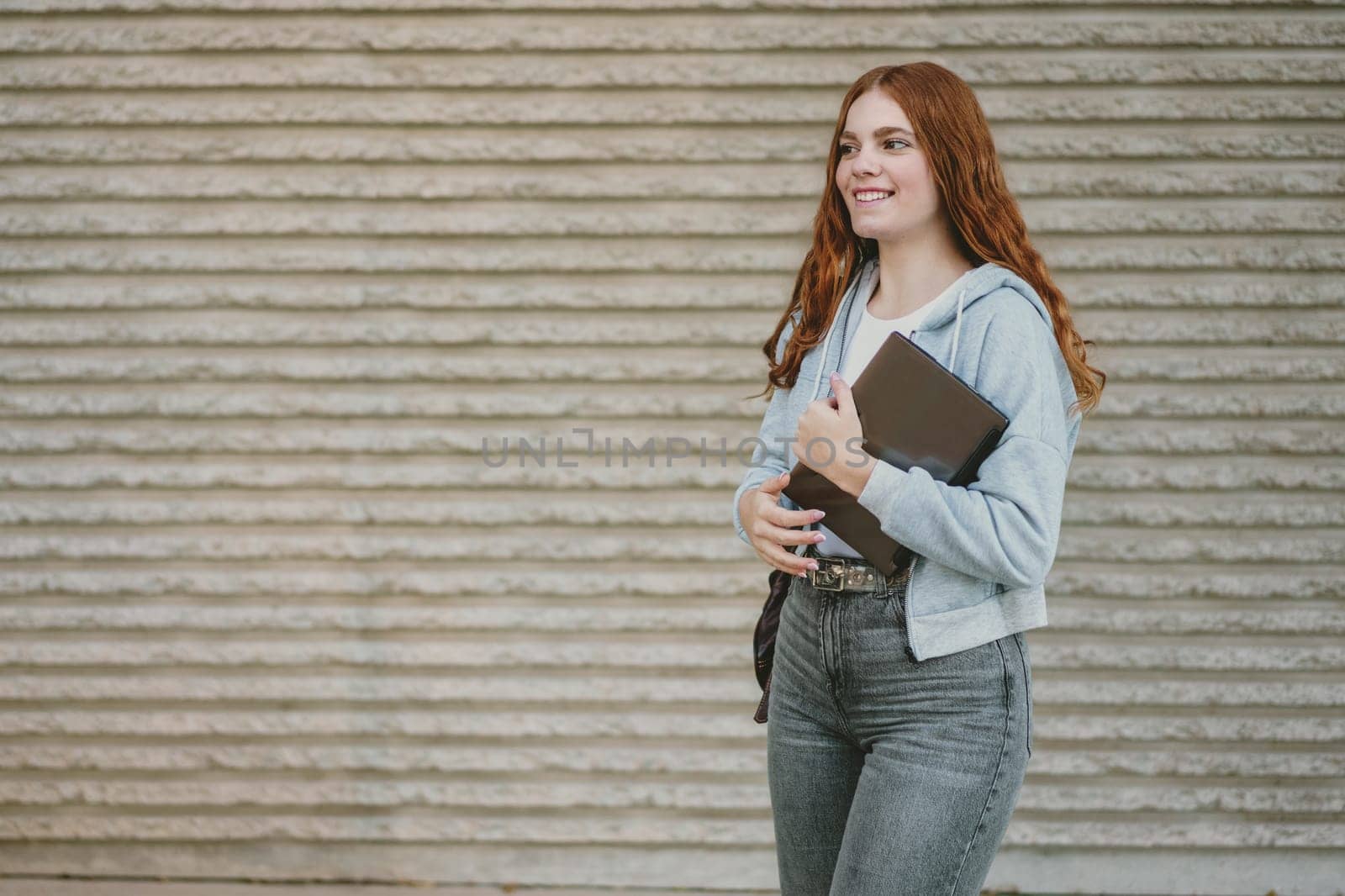 A stylish and fashionable young woman with beautiful curly red hair is smiling brightly while walking outdoors, holding a notebook, which radiates her confidence and charm effortlessly