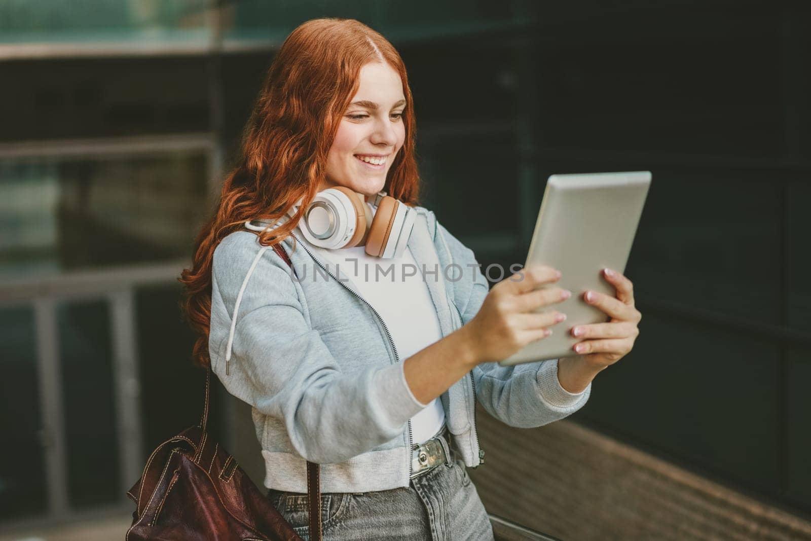 A cheerful and lively teenage girl with vibrant red hair is using a tablet to engage in a fun video call while wearing stylish headphones in a bustling urban environment, surrounded by city life