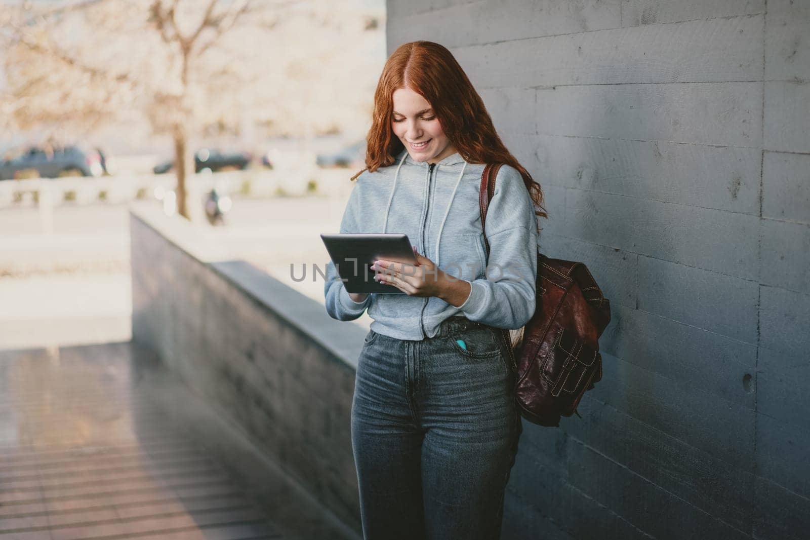 A young woman with vibrant red hair stands confidently against a concrete wall, fully engaged with her tablet in a lively outdoor urban setting, showcasing modern technology and a sense of leisure