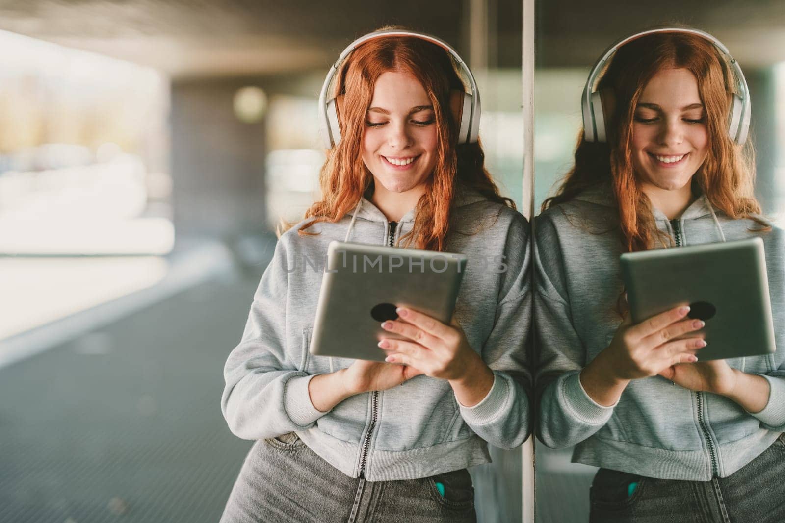 In a lively urban setting, a cheerful young woman with curly red hair finds joy through music, engaging with her tablet and wearing stylish headphones, fully immersed in her entertainment