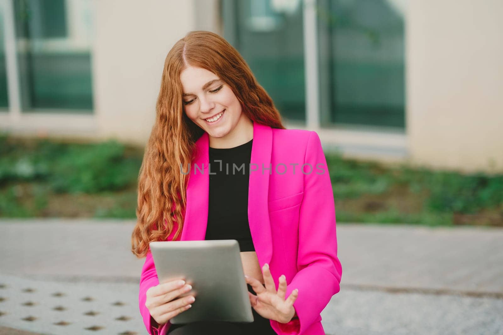 A cheerful young woman in a pink blazer is enjoying her outdoor leisure time. She holds a tablet while appreciating the fresh air, bright sunlight, and beautiful greenery around her
