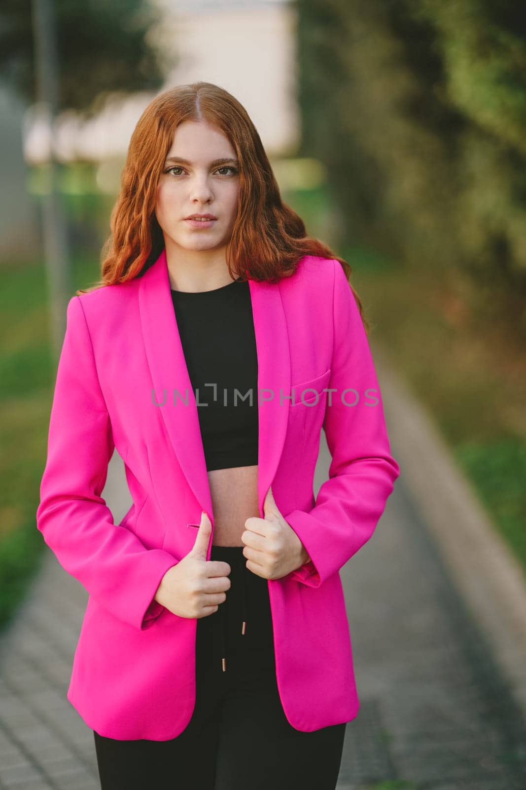 A stylish young woman confidently poses in a vibrant pink blazer and trendy black crop top, set against a beautifully blurred outdoor background that highlights her flair for modern fashion