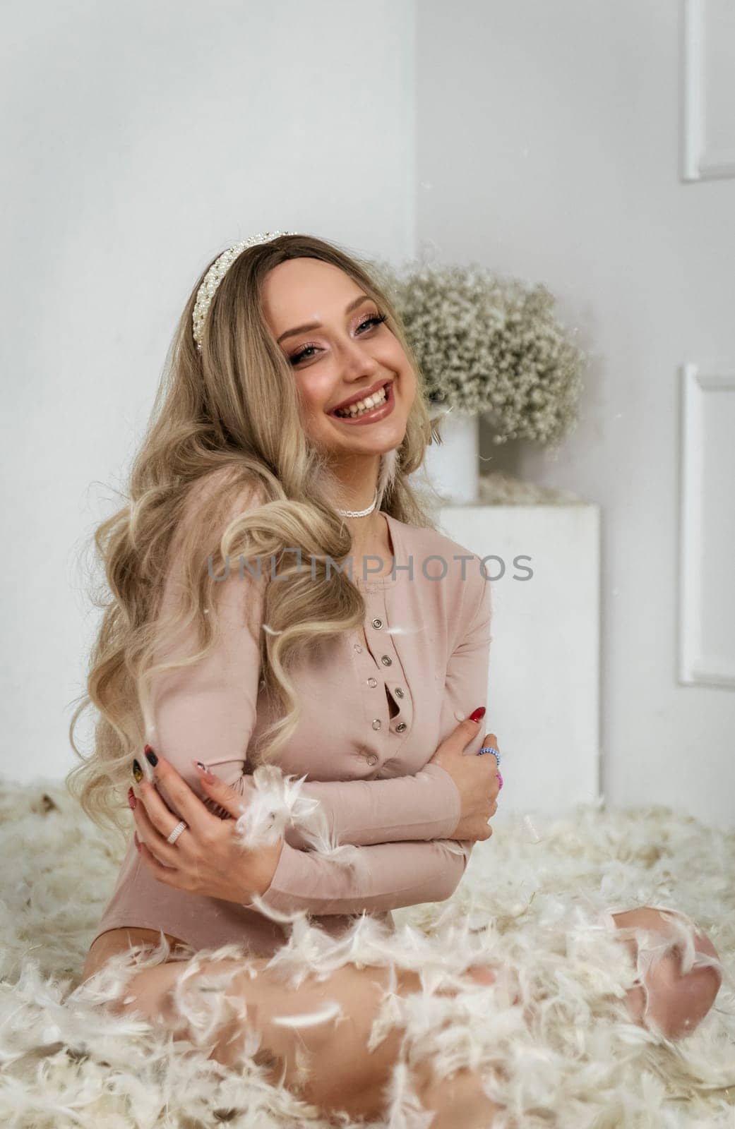 young beautiful blonde girl among feathers. feathers fly in the air. the girl has light long hair she smiles a beautiful smile. on her neck she has pearls on her head crown