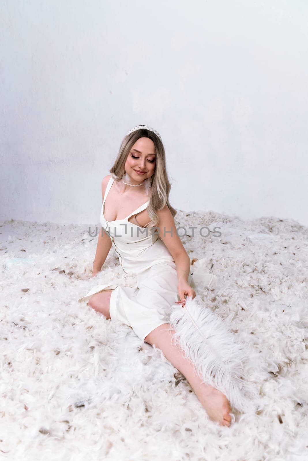 a very beautiful blonde is sitting in a white room with feathers flying in it.the girl has long hair. she is wearing a white silk dress. she is holding a large feather in her hand and touching her leg