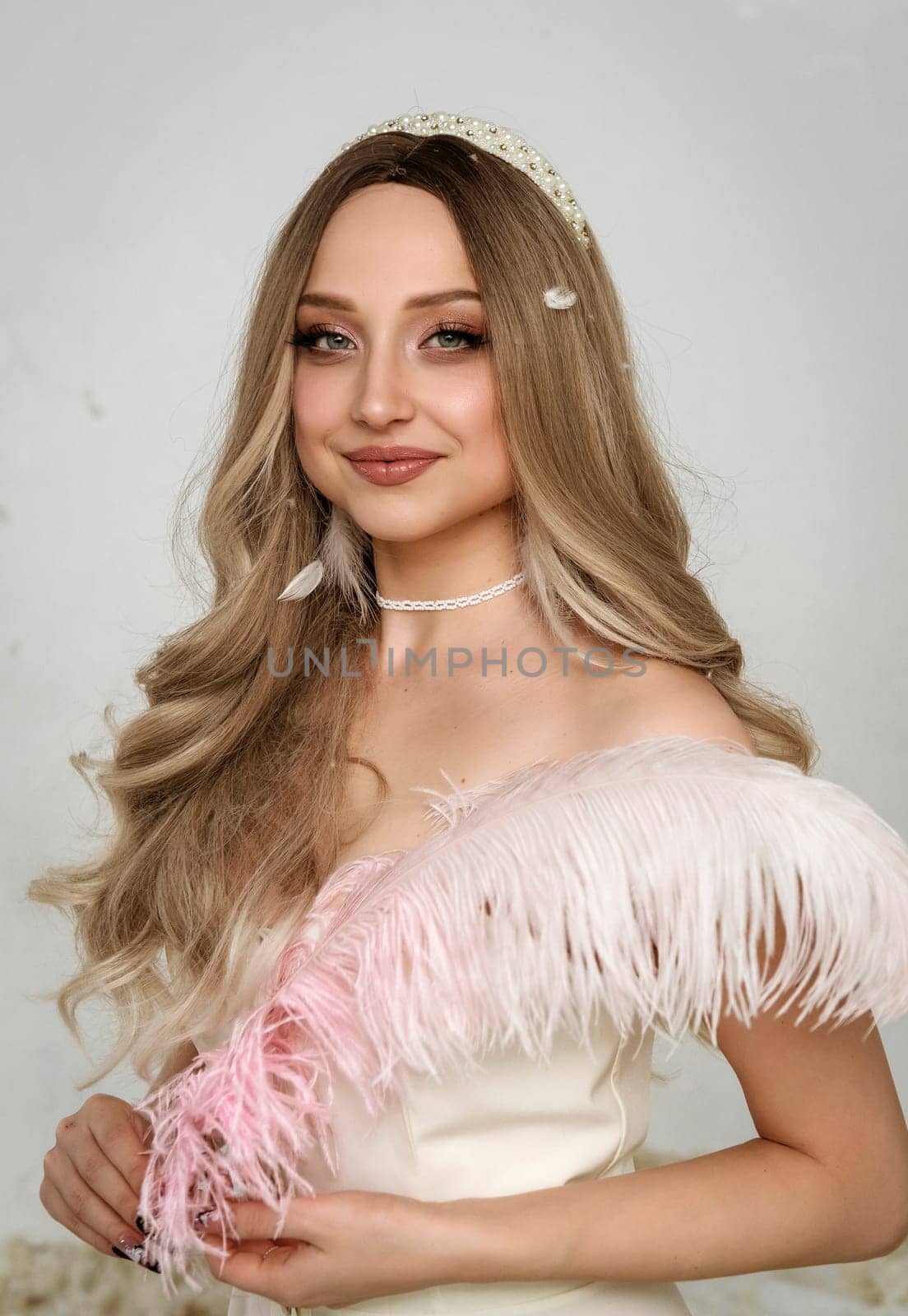 Portrait of a young blonde girl holding a pink feather. The girl smiles and looks at the camera. She has beautiful clear skin and an open smile. Her hair is styled in a wave. she has pearls on her neck