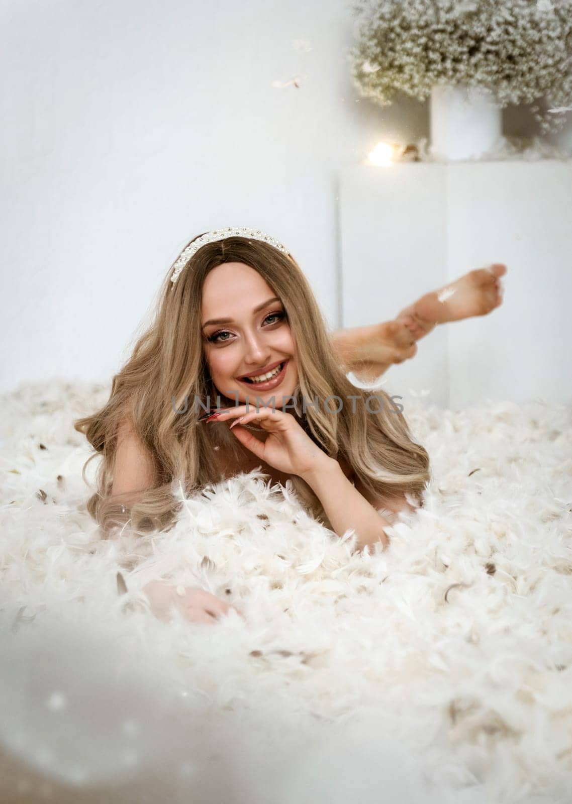 young beautiful blonde girl lies among feathers. feathers fly through the air in a white room. girl looks at the camera and smiles a happy smile