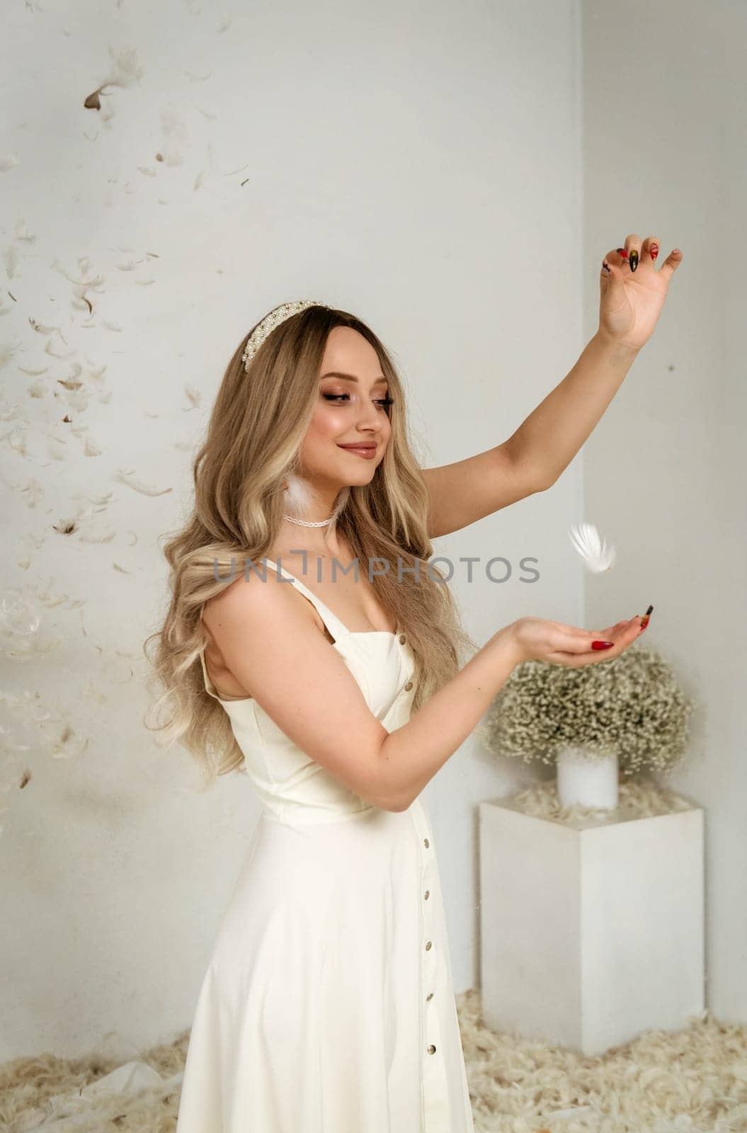 A young beautiful blonde girl stands among feathers. Feathers fly through the air in a white room. One feather flies between her hands. There is a vase with white flowers in the room.