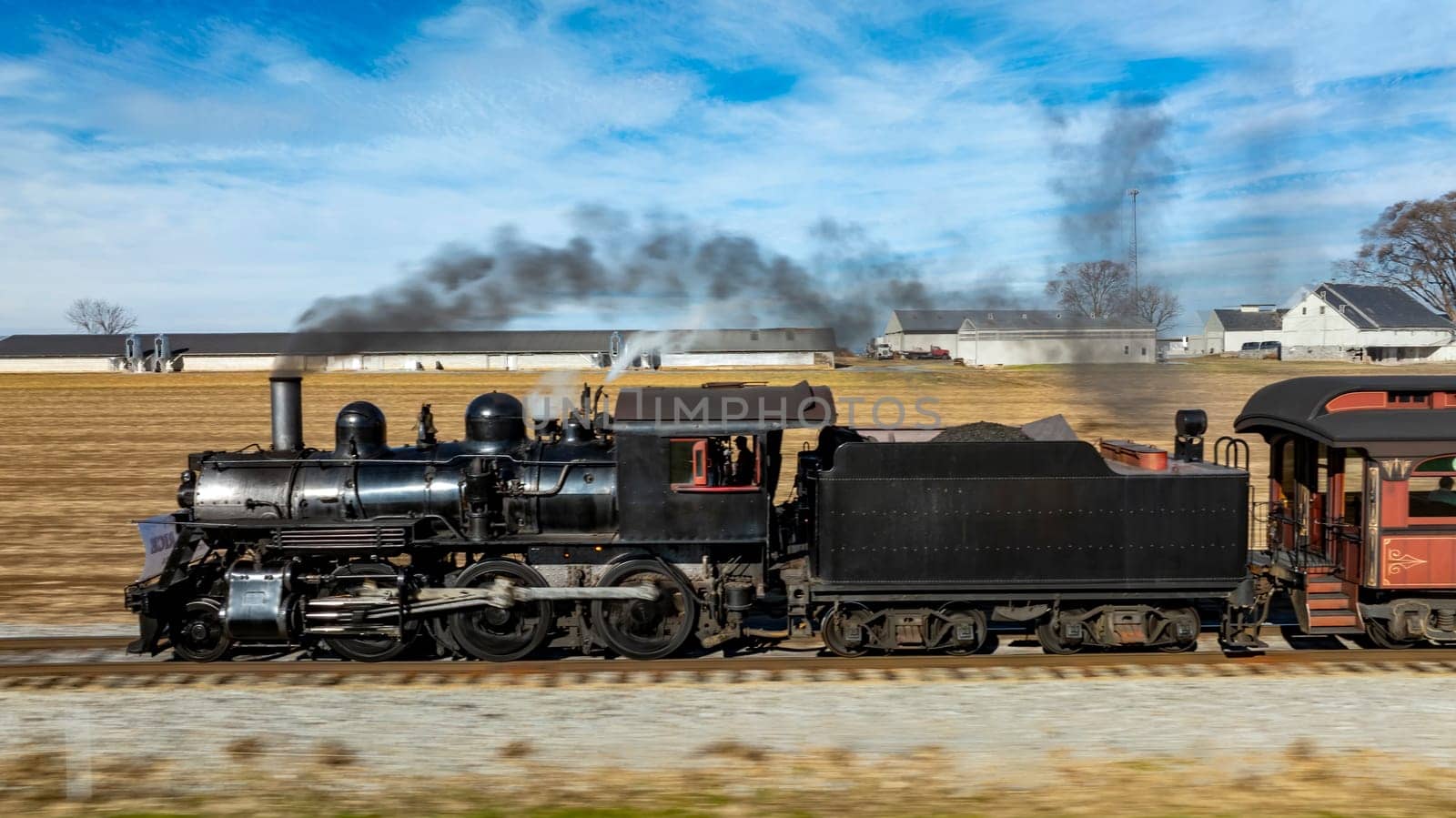 A vintage steam locomotive powers along the tracks, sending plumes of smoke into the blue sky. Surrounding fields and farmhouses enhance the picturesque countryside setting.