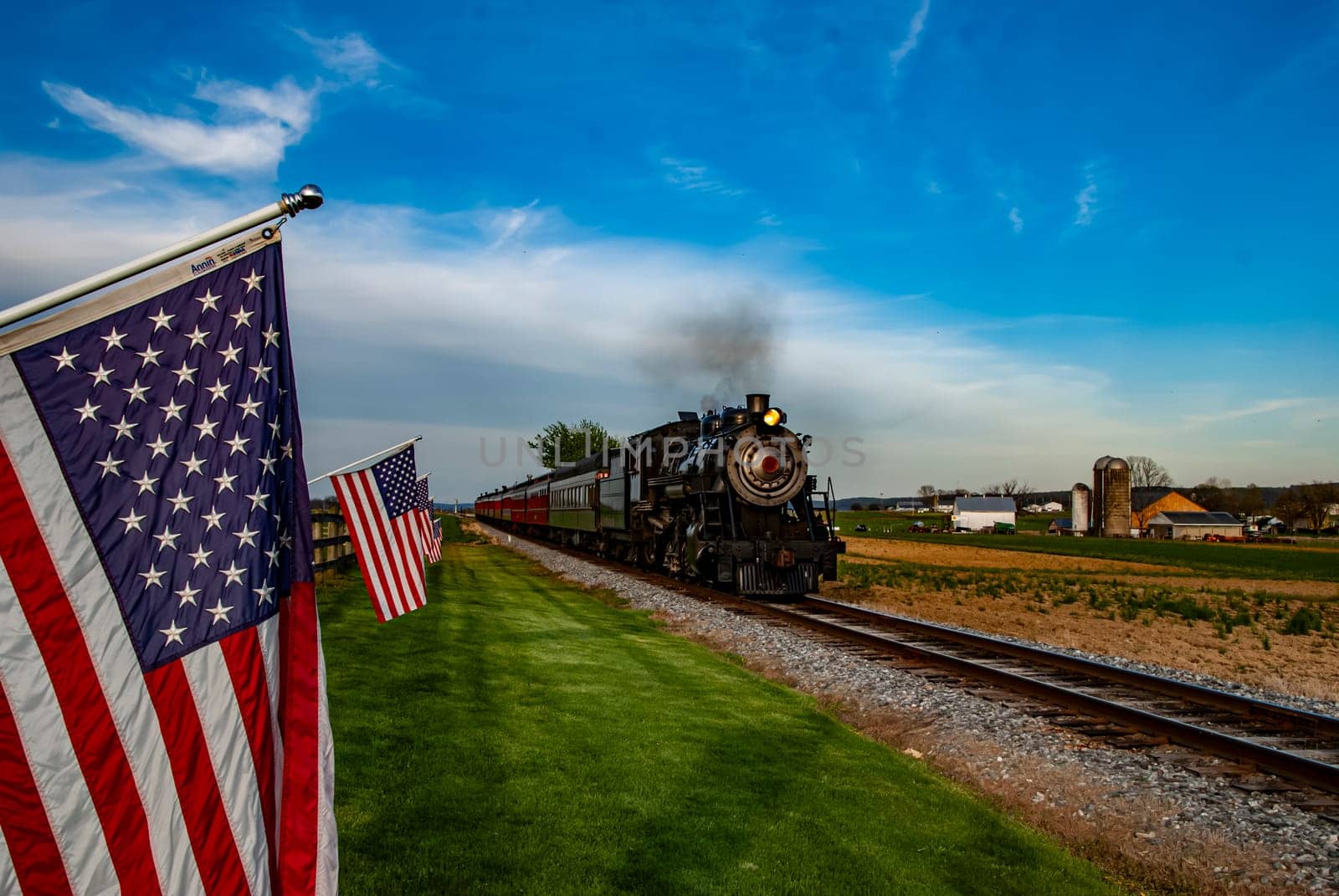 A vintage steam train chugs along scenic tracks bordered by green fields and American flags. The atmosphere is bright and inviting during a clear afternoon with a blue sky.