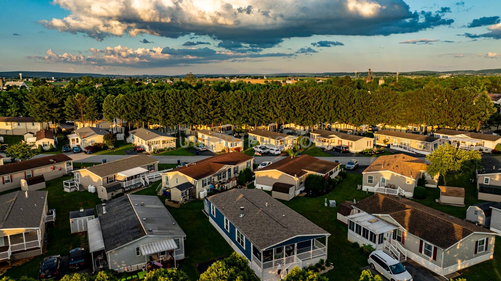 Aerial view captures a Mobile, Prefab, Manufactured Home neighborhood with houses surrounded by tall trees. The sun sets in the background, casting a warm glow over the rooftops lush landscape.