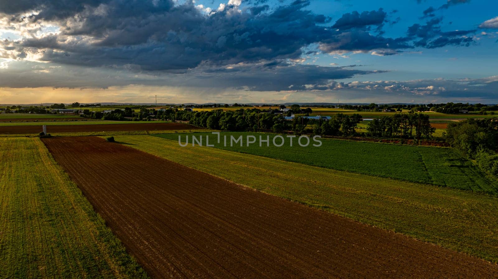 As the sun sets in a vibrant sky, vast agricultural fields stretch across the landscape, dotted with trees and farm structures, creating a serene and picturesque rural scene.