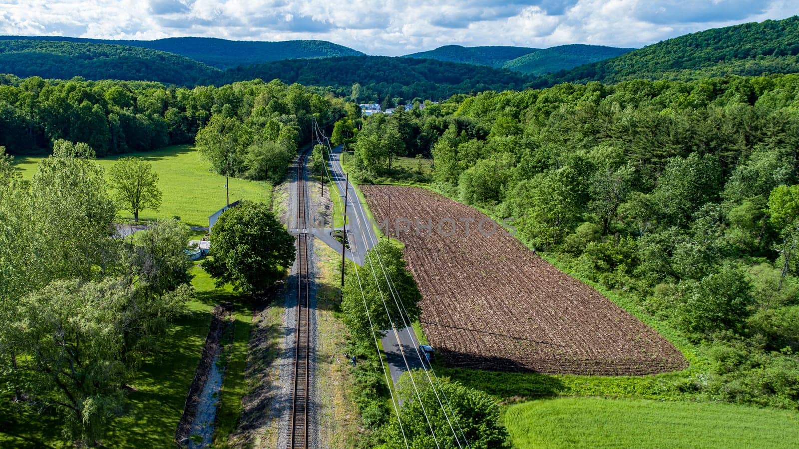 A peaceful countryside landscape features train tracks running alongside a tilled field, surrounded by greenery and rolling hills under a bright blue sky with fluffy clouds.