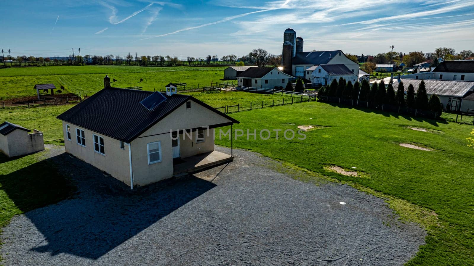 An Amish one room school house sits on a gravel area, surrounded by expansive green fields, barns, and distant structures under a bright blue sky.