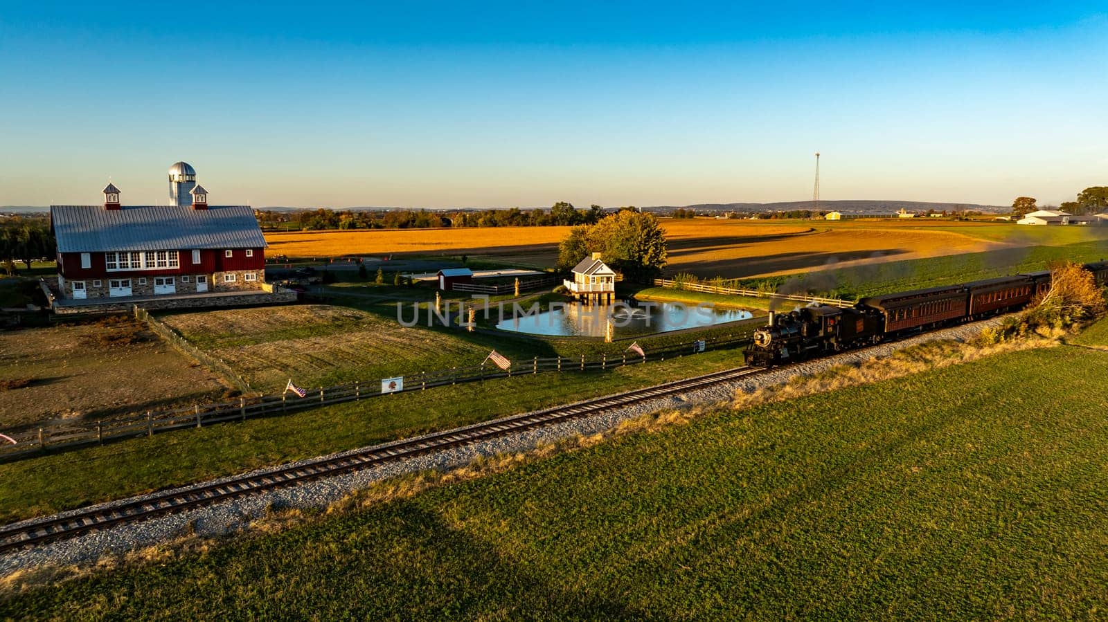 Ronks, Pennsylvania, USA, October 20 2024 - Historic steam train moves along the tracks, passing a picturesque red barn expansive farmland. The sun sets, casting warm golden hues across the landscape,