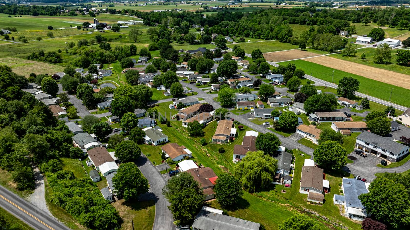 Aerial view of a tranquil rural community featuring various Mobile, Prefab, Manufactured houses situated near lush green fields and trees. The landscape shows harmonious blend of residences farmland.