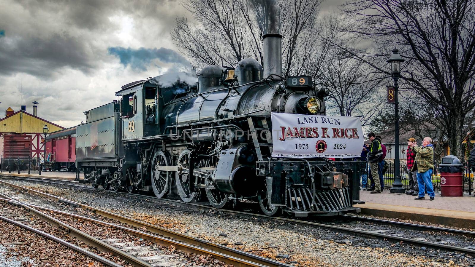 Strasburg, Pennsylvania, USA, December 31, 2024 - A steam locomotive, adorned with a farewell banner, stands at a bustling railway station. Rail enthusiasts gather to witness its last journey, creating a nostalgic atmosphere.