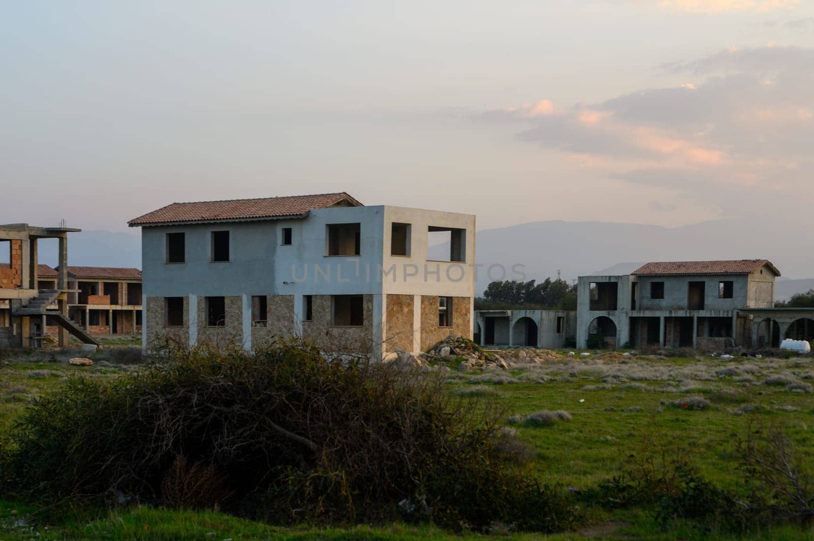 Ruins of unfinished constructions stand silently as the sun sets, casting shadows across the overgrown landscape.