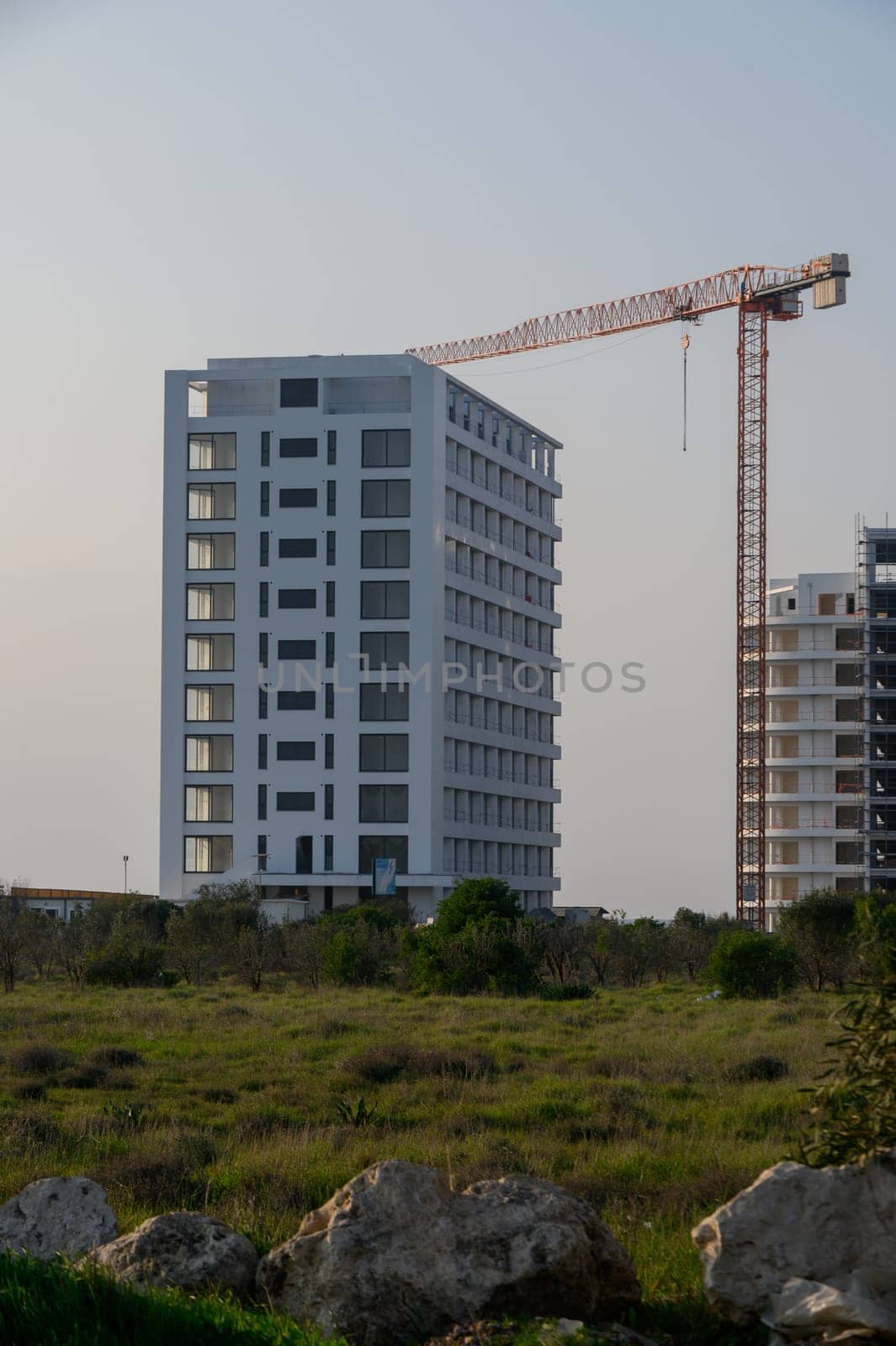 A modern building rises beside a construction crane, surrounded by lush greenery under a soft evening light.