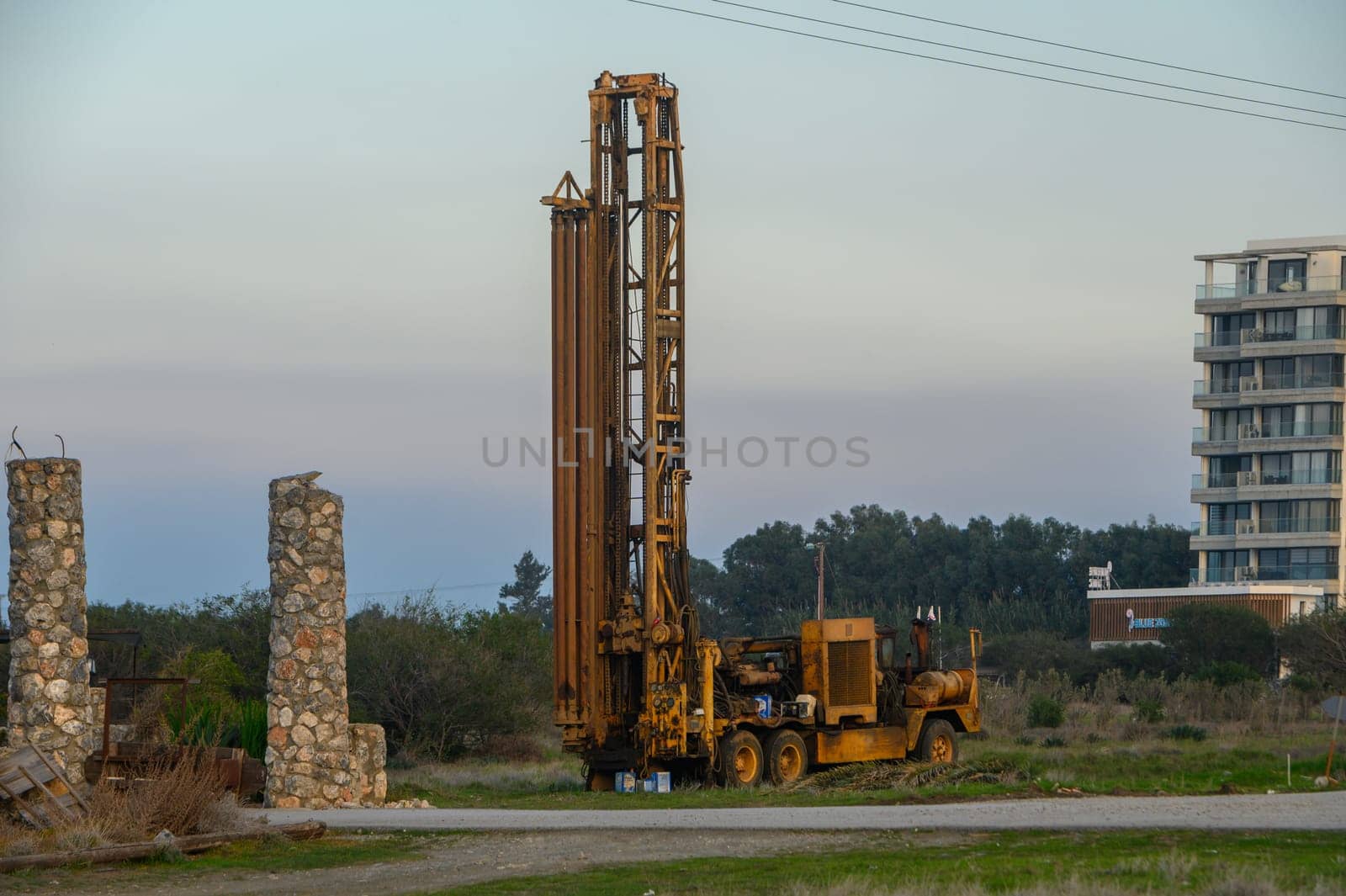 A towering drilling rig stands amidst a transitioning landscape under a soft afternoon sky.