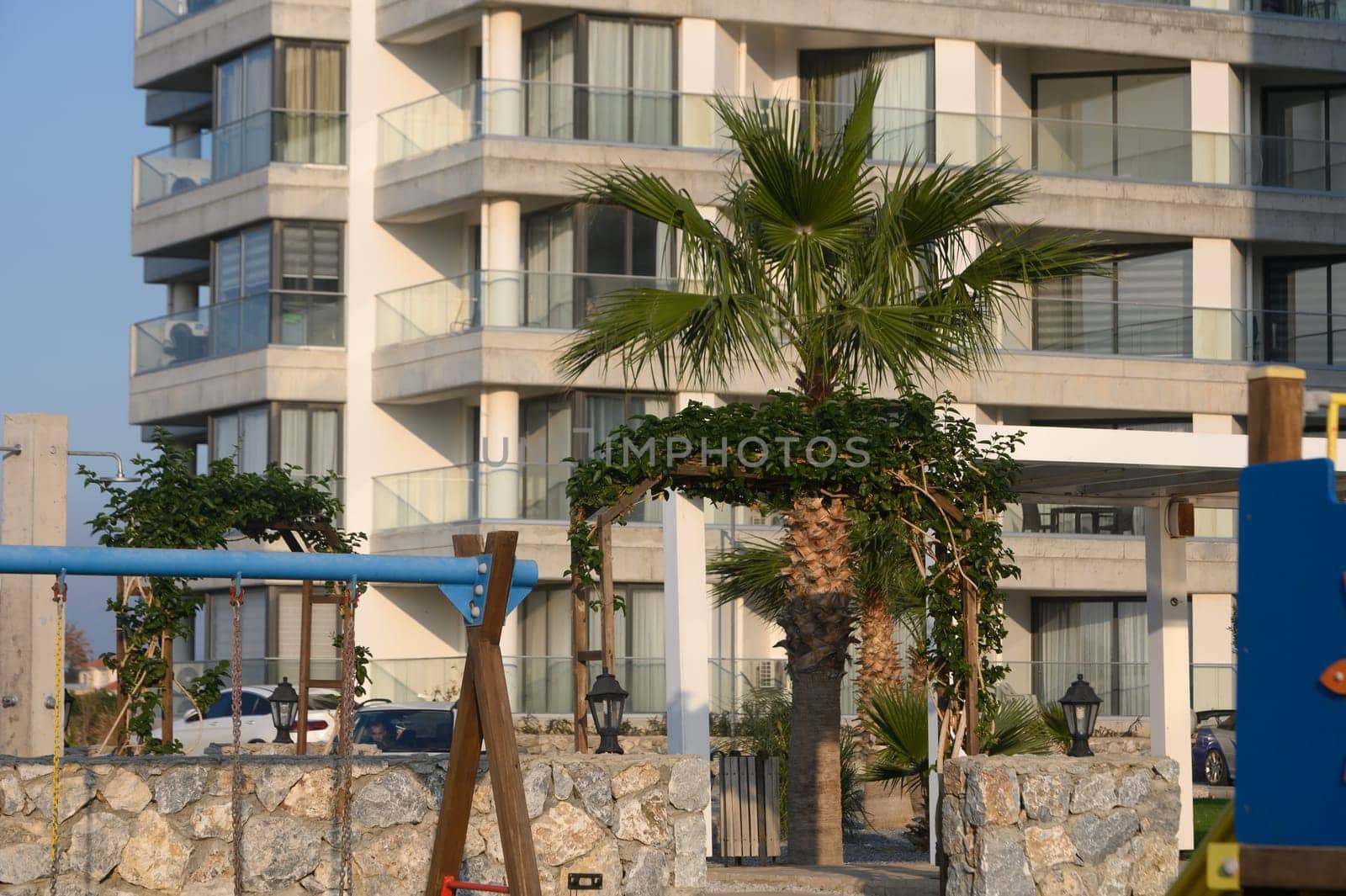 Palm trees sway gently by a modern building, overlooking a tranquil playground while the sun sets on the horizon.