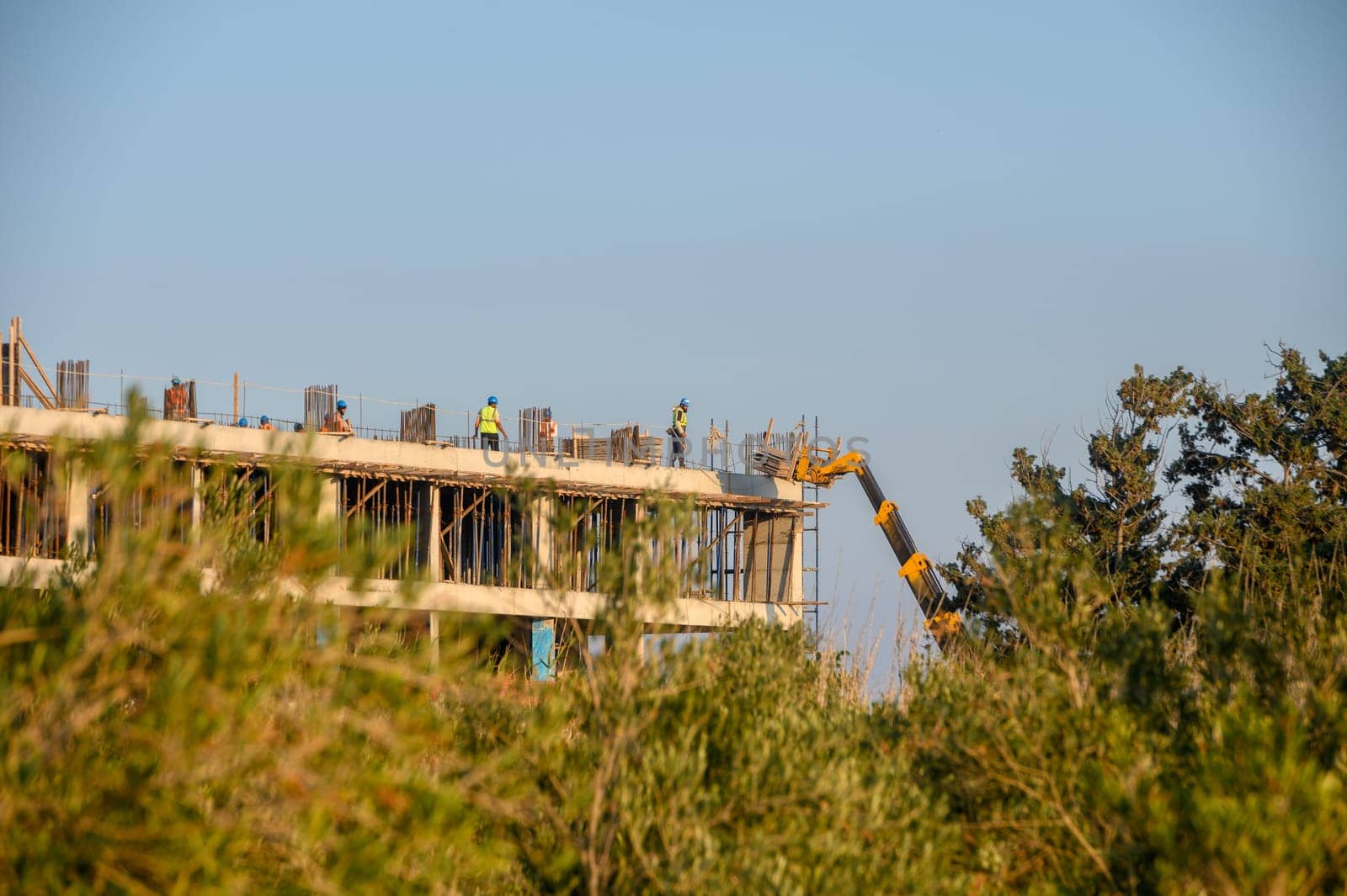 Workers collaborate on a construction site, raising a new structure amidst greenery under the warm sun.
