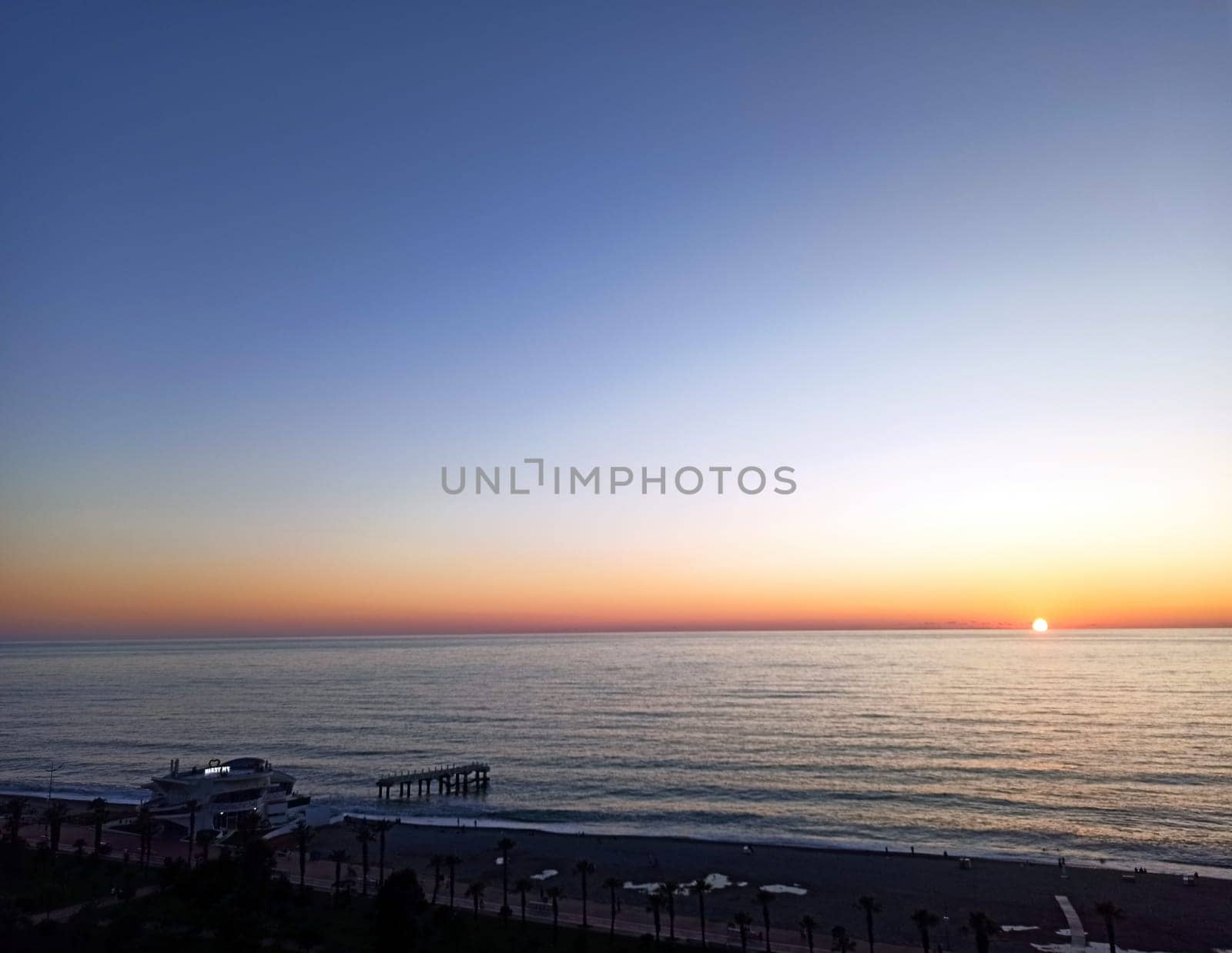 The sun sets on the horizon, casting warm hues across the tranquil ocean waves. A pier stands in the distance, silhouetted against the colorful sky, creating a serene atmosphere.