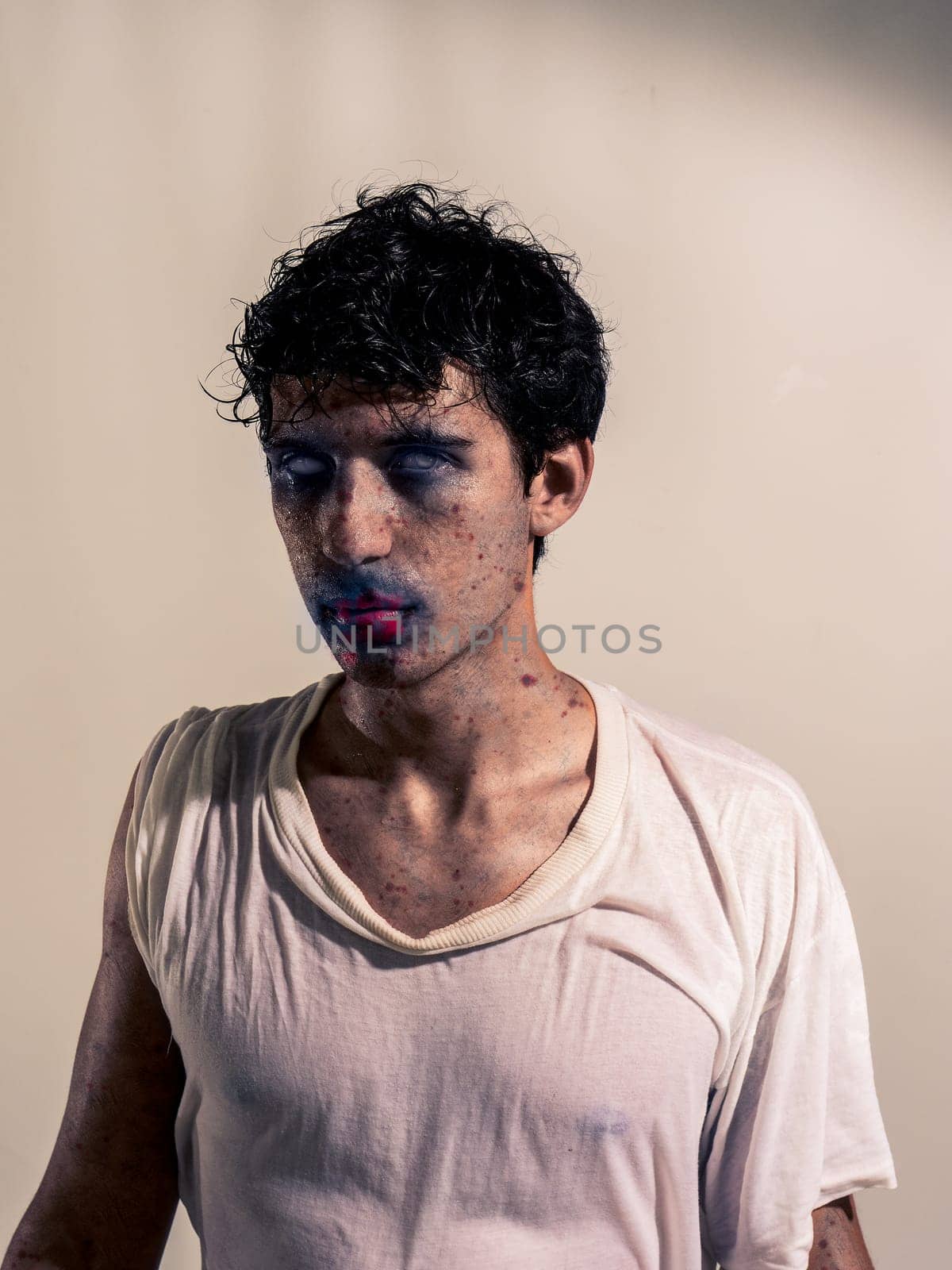 Scary male zombie standing on dark grey background, with white eyes and bloody mouth, looking at camera
