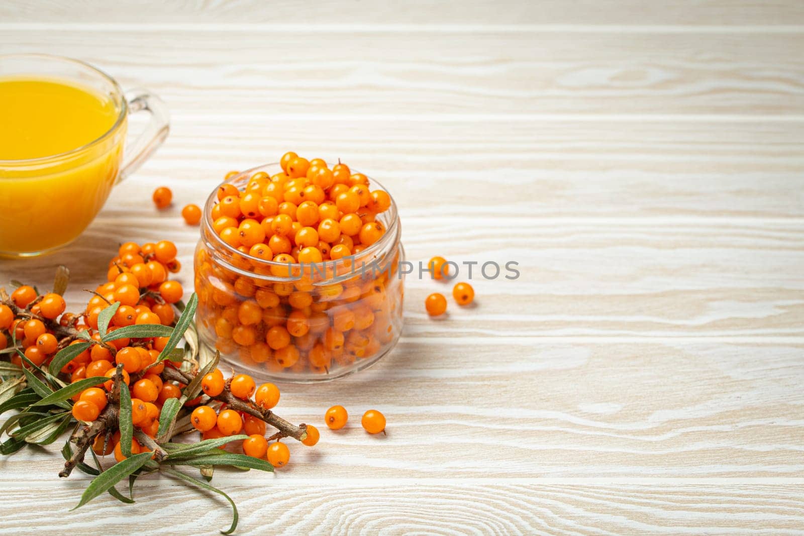 Sea buckthorn healthy hot tea in transparent glass cup, ripe berries in glass jar and branches with leaves on white wooden rustic background, for skin, heart, vessels and immune system. Copy space.