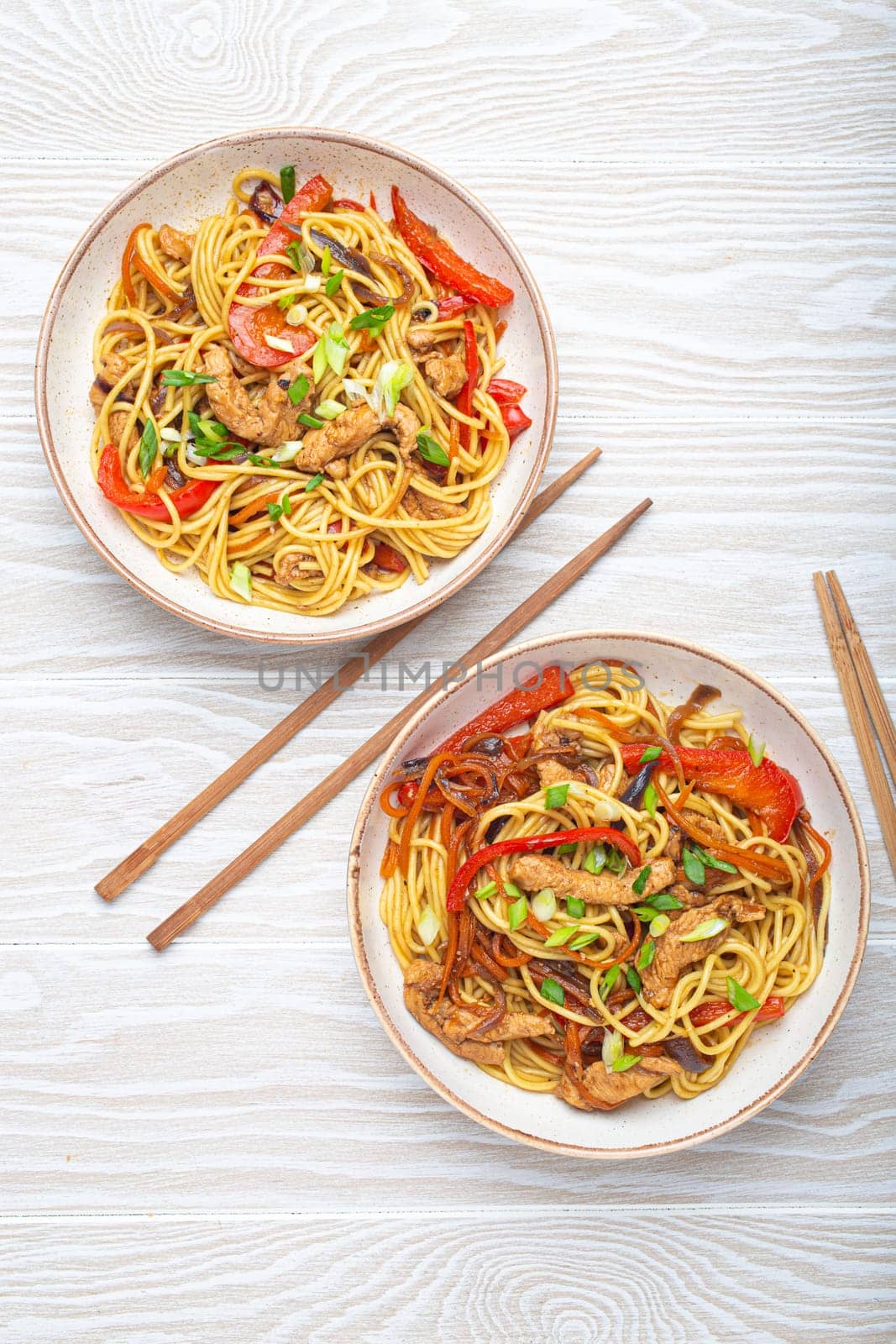 Two bowls with Chow Mein or Lo Mein, traditional Chinese stir fry noodles with meat and vegetables, served with chopsticks top view on rustic white wooden background table.