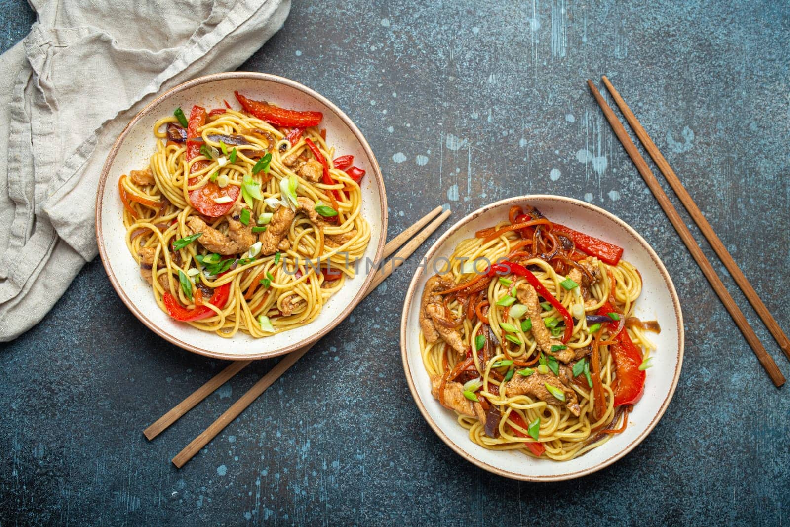 Two bowls with Chow Mein or Lo Mein, traditional Chinese stir fry noodles with meat and vegetables, served with chopsticks top view on rustic blue concrete background.