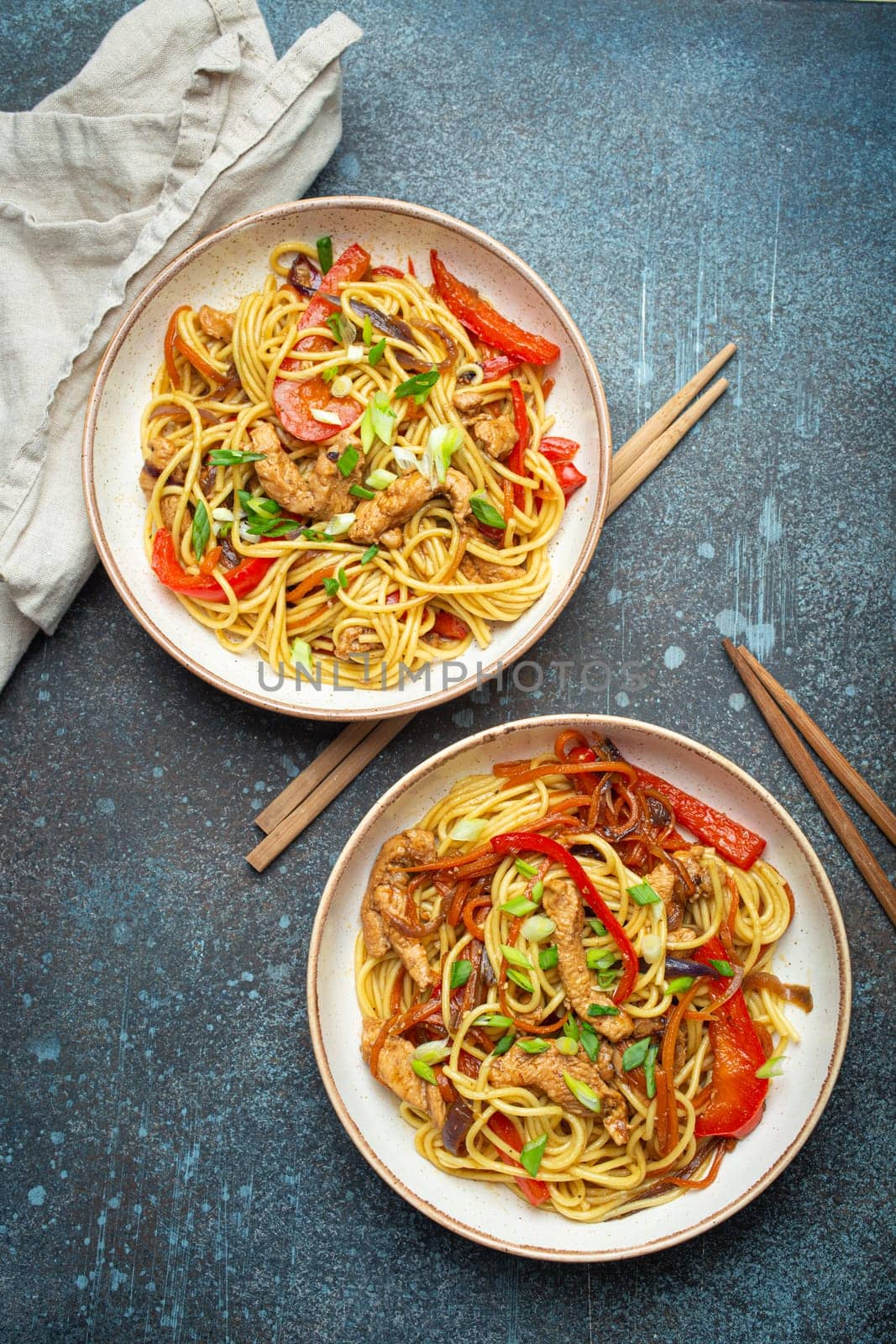 Two bowls with Chow Mein or Lo Mein, traditional Chinese stir fry noodles with meat and vegetables, served with chopsticks top view on rustic blue concrete background.