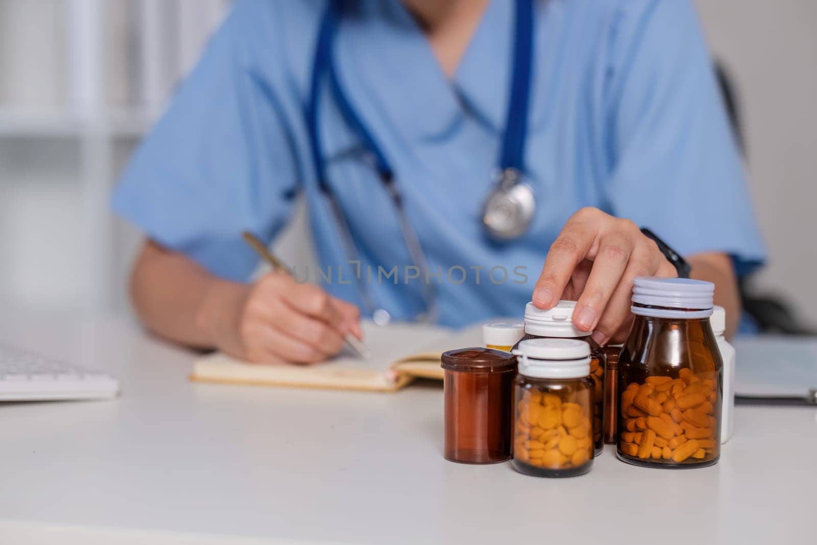 Asian female doctor, GP therapist holding pill bottle in hand, writing a prescription, sitting at prescription table, pharmaceutical concept. Closeup view. by wichayada