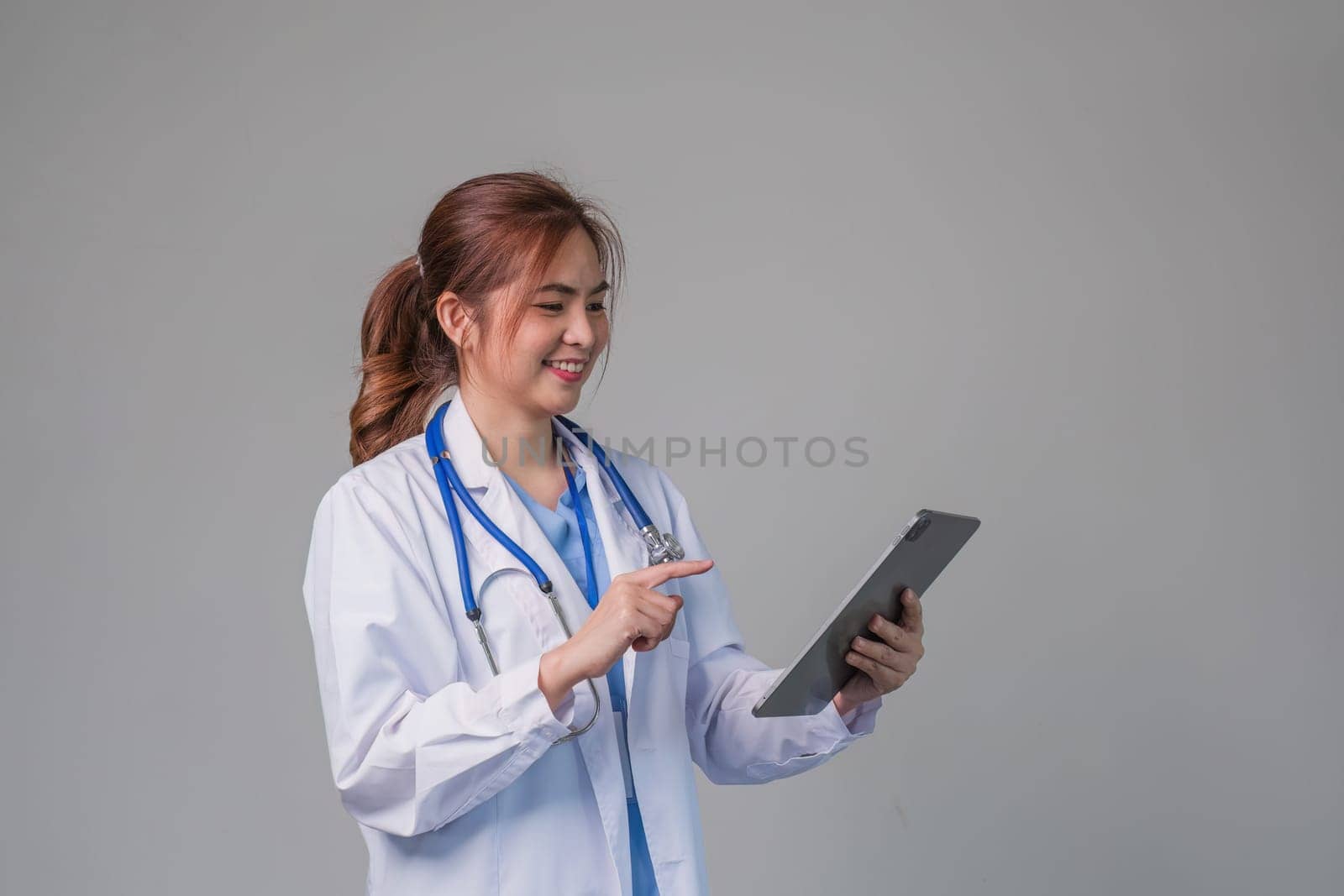 Portrait of smiling female doctor holding digital tablet wearing medical robe and stethoscope, grey background..