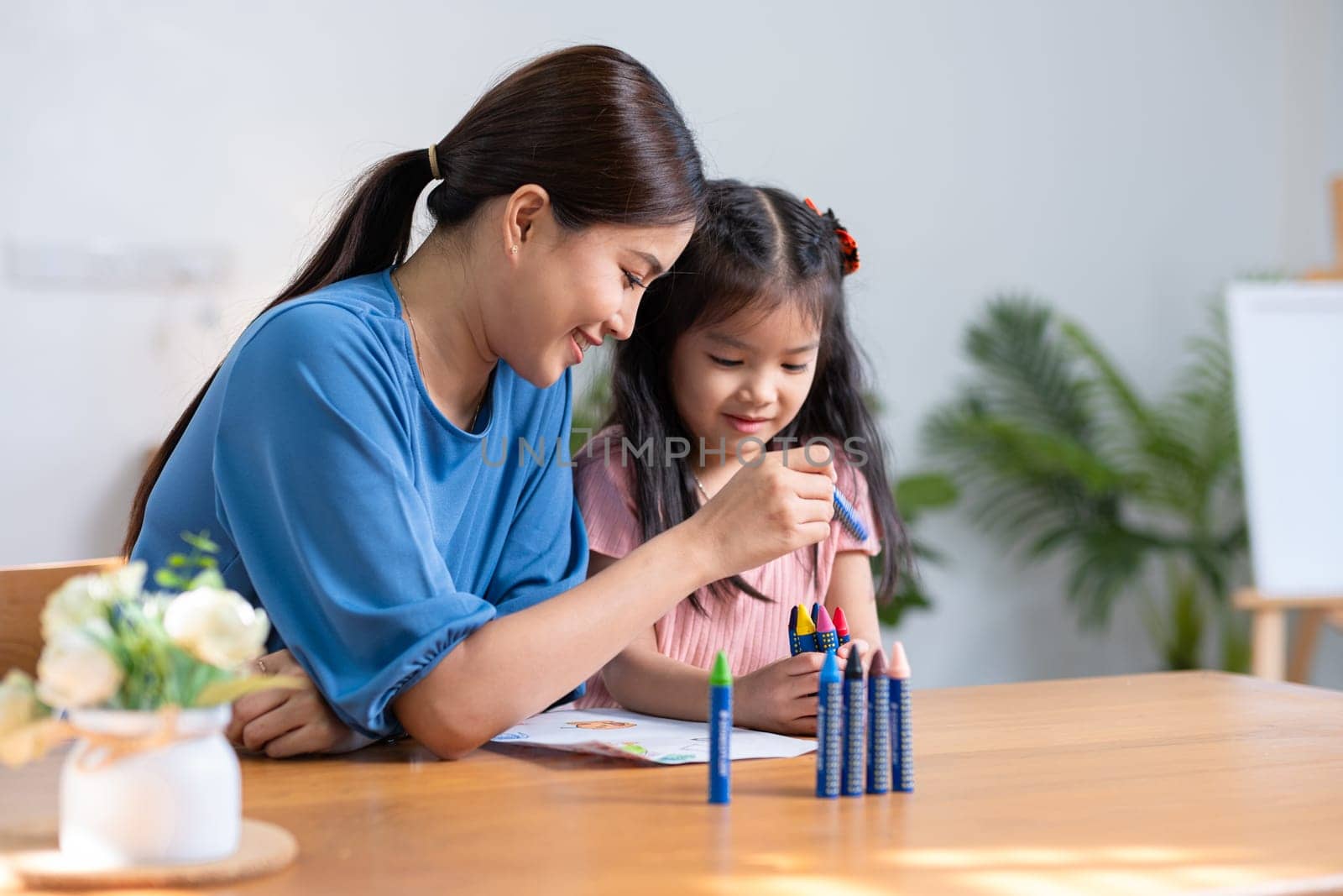 A young family spends their free time together in the living room at home. Mother and little daughter draw pictures with crayons on paper, smiling happy..