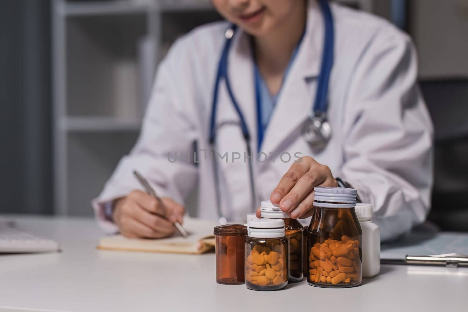 Asian female medic doctor gp therapist wear white coat holding pills bottle in hand writing medical prescription sitting at work desk prescribing pharmacy medicine concept. Closeup view by wichayada