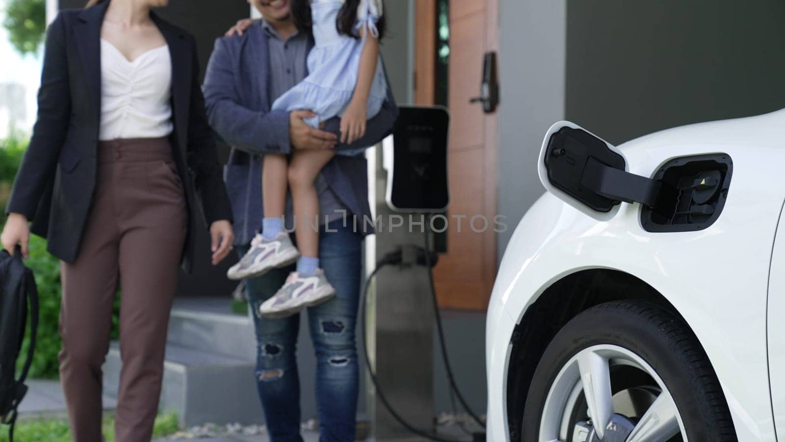 Progressive young parents and daughter living in a home with an electric vehicle and electronic vehicle charging station. Green and clean energy from electric vehicles for healthy environment.