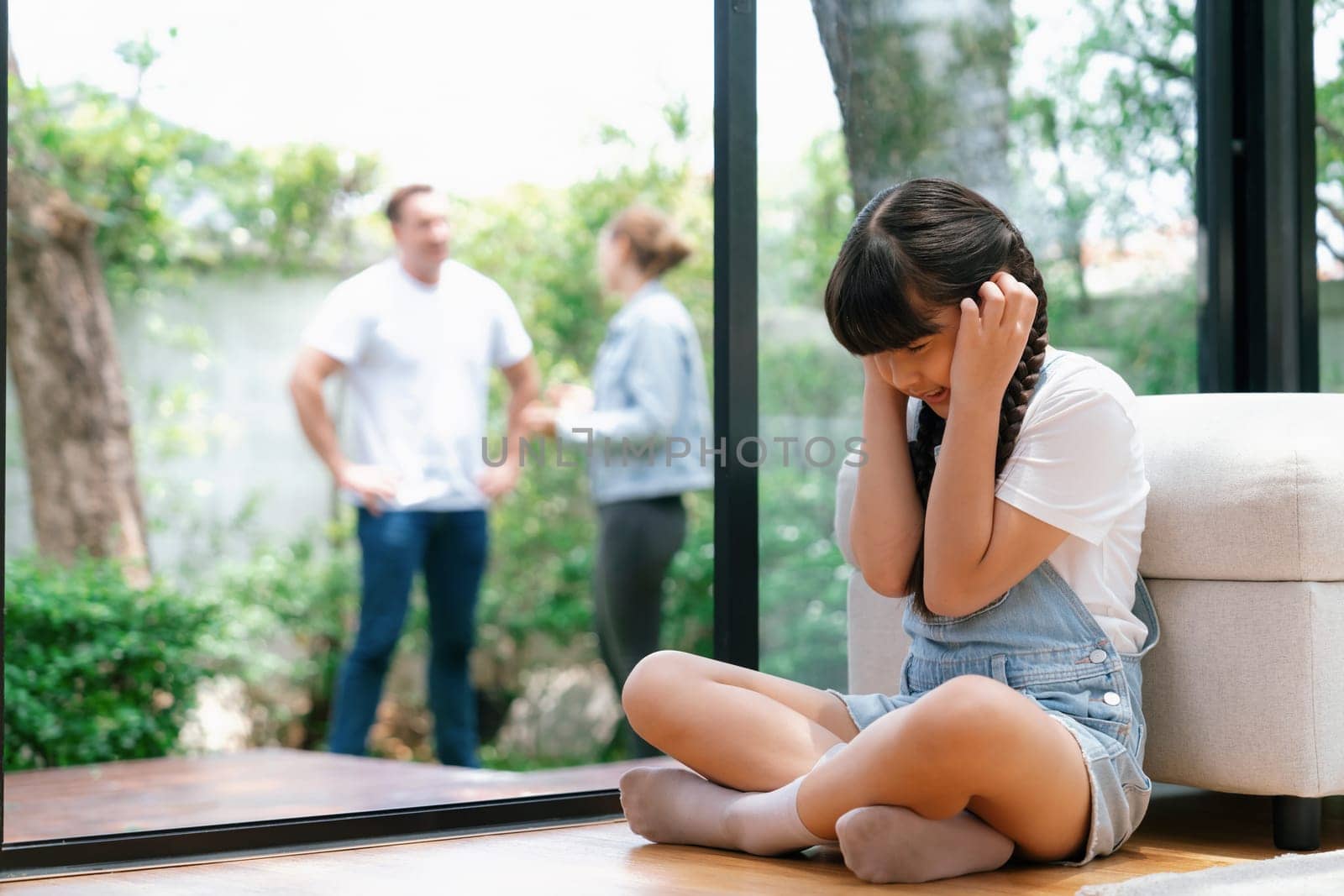 Stressed and unhappy young girl huddle in corner, cover her ears blocking sound of her parent arguing in background. Domestic violence at home and traumatic childhood develop to depression. Synchronos