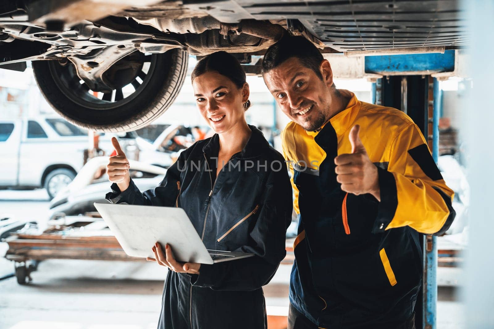 Two vehicle mechanic working together underneath lifted car, conduct car inspection with laptop. Automotive service technician in uniform carefully make diagnostic troubleshooting. Oxus