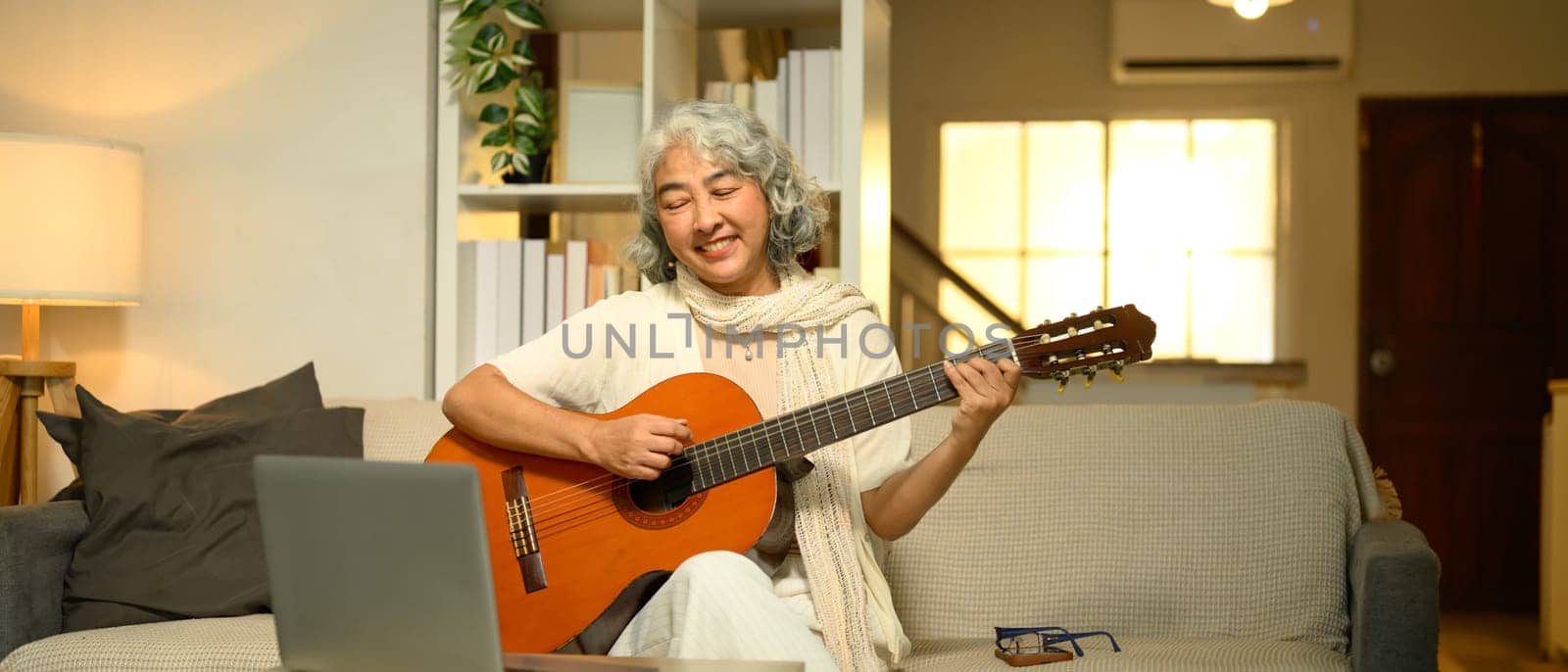 Cheerful elderly woman plays an acoustic guitar while learning to play acoustic guitar online.