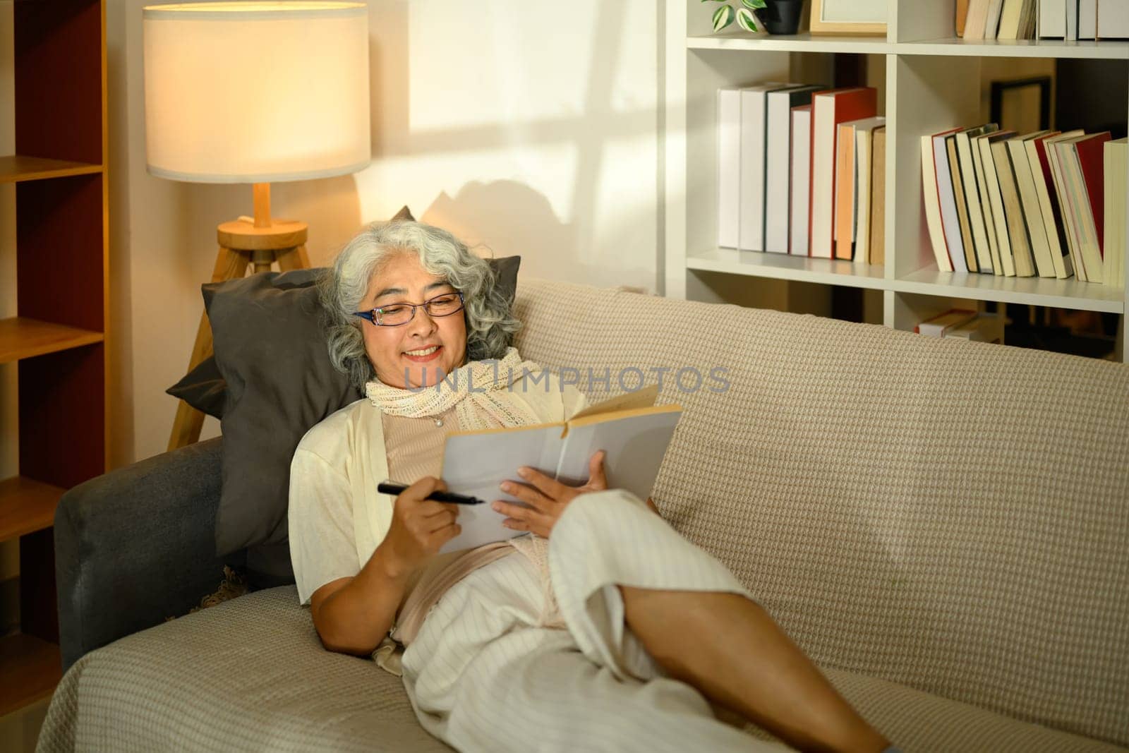 Happy elderly woman with gray hair sitting on sofa and writing in notebook with a smile.