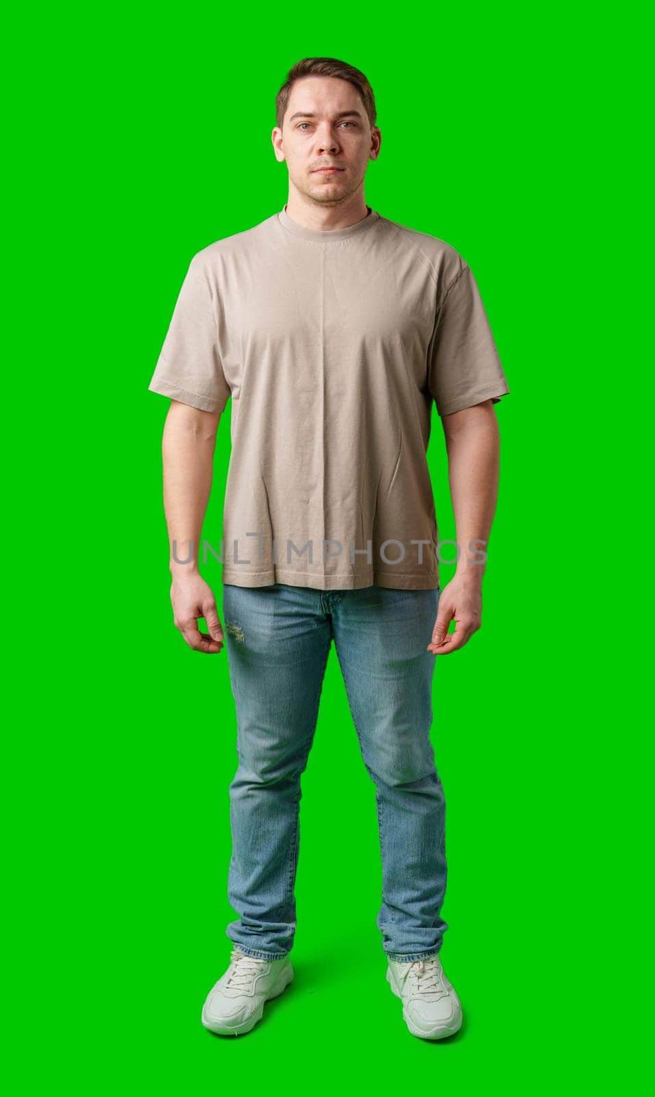 Young man stands confidently in a plain beige t-shirt and jeans against a bright green background in studio