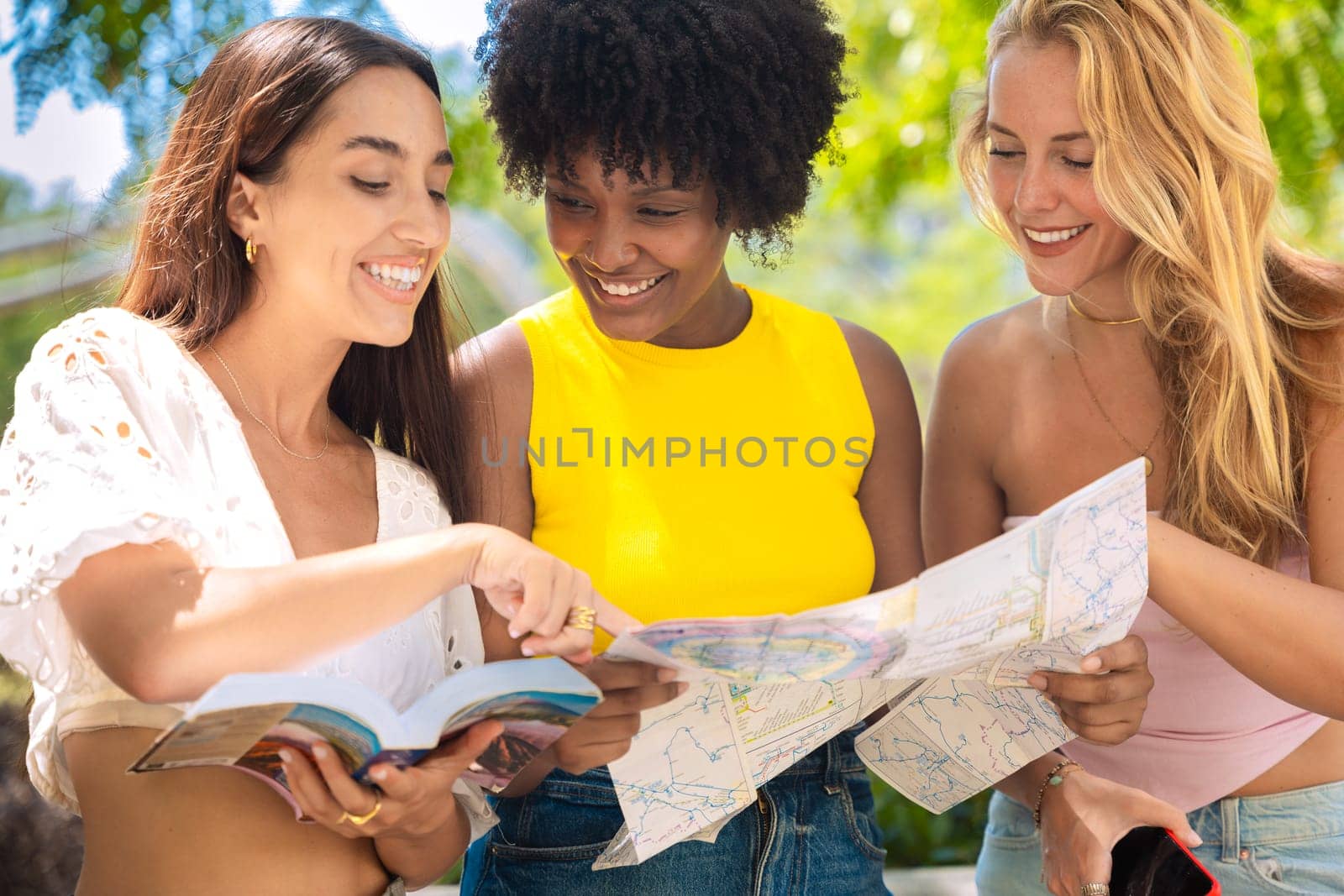 Three happy young women stand outdoors, looking at maps and a travel guide. They smile while planning their trip, enjoying a fun adventure in a sunny urban setting with trees and buildings.
