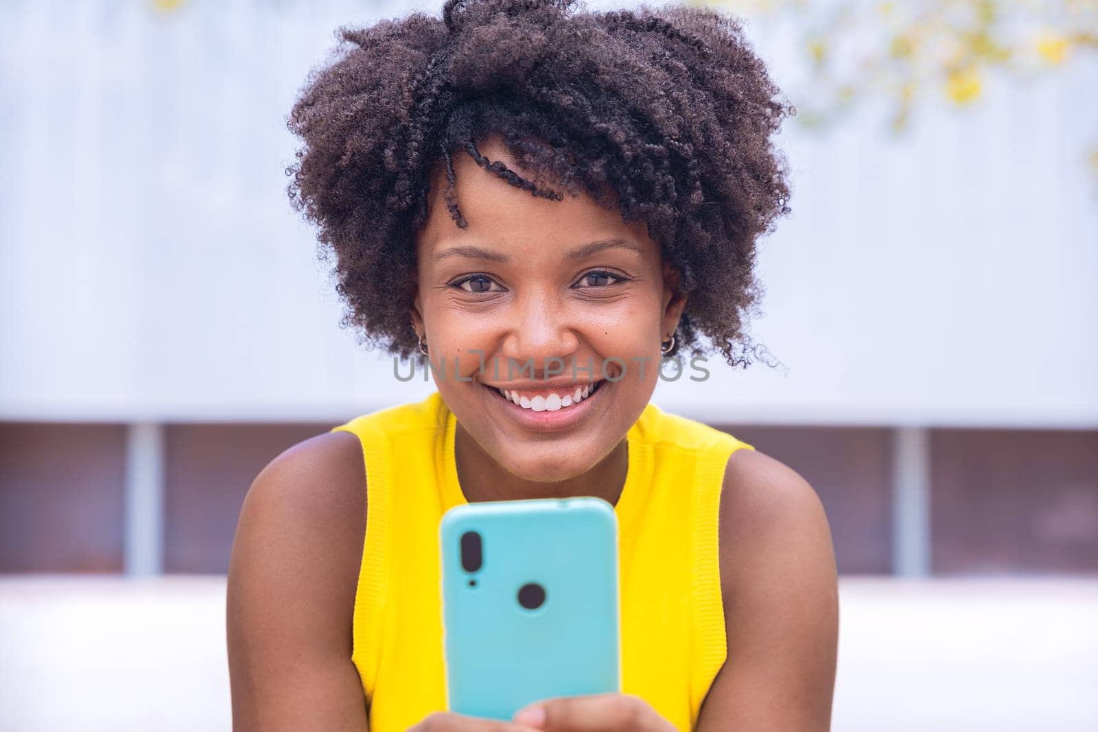 A young woman with natural curly hair wearing a yellow top rests her face on her hand while holding a smartphone. Calm and happy moment captured in a bright outdoor setting.