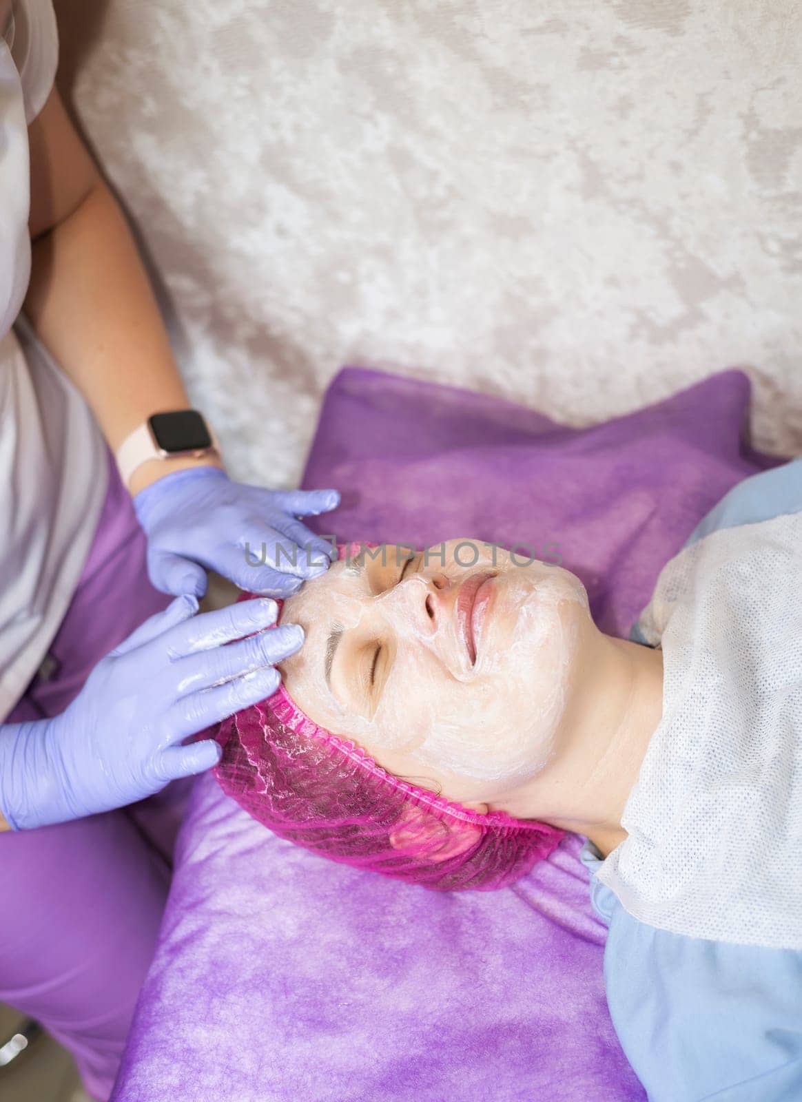 A cosmetologist is performing a facial procedure on a client. The doctor is wearing purple rubber gloves. The cosmetologist is applying cream to a female client's face.