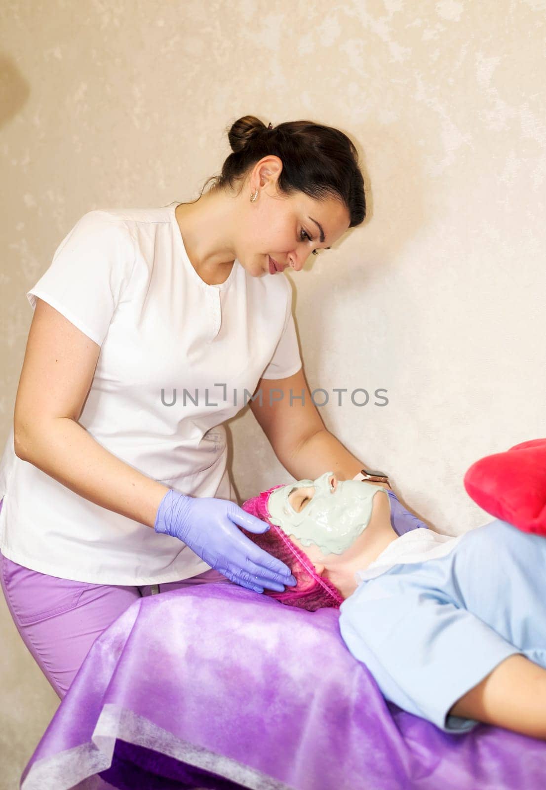 A female cosmetologist doctor makes a facial mask for a client. A female client lies on a couch. The cosmetologist removes an alginate mask from her face. facial rejuvenation mask in facial clinic