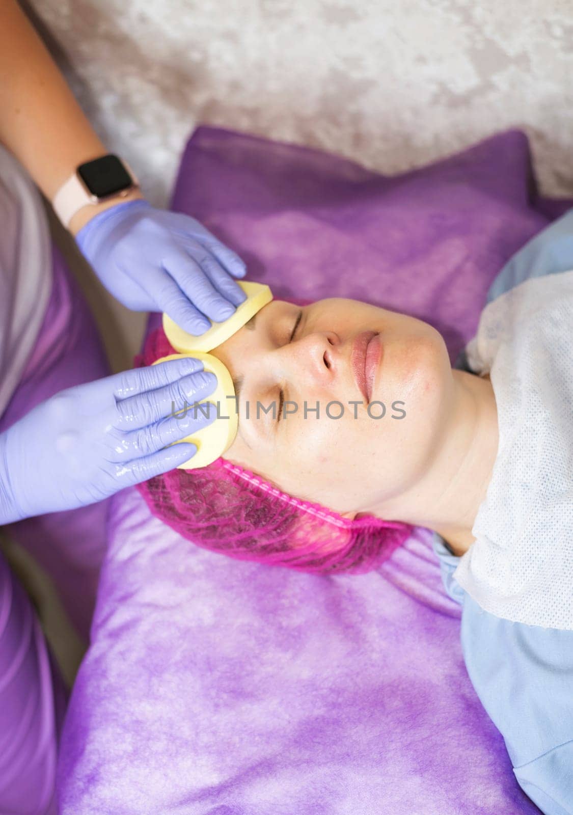 A cosmetologist wipes a woman client's face with sponges. The cosmetologist holds two sponges. The client lies on a couch, her hair is tucked into a cap. The colors in the photo are blue and purple