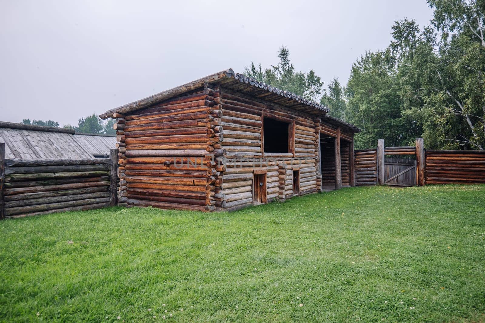 Weathered wooden barn nestled in verdant meadow, surrounded by rustic fence, capturing rural russian landscape's timeless charm