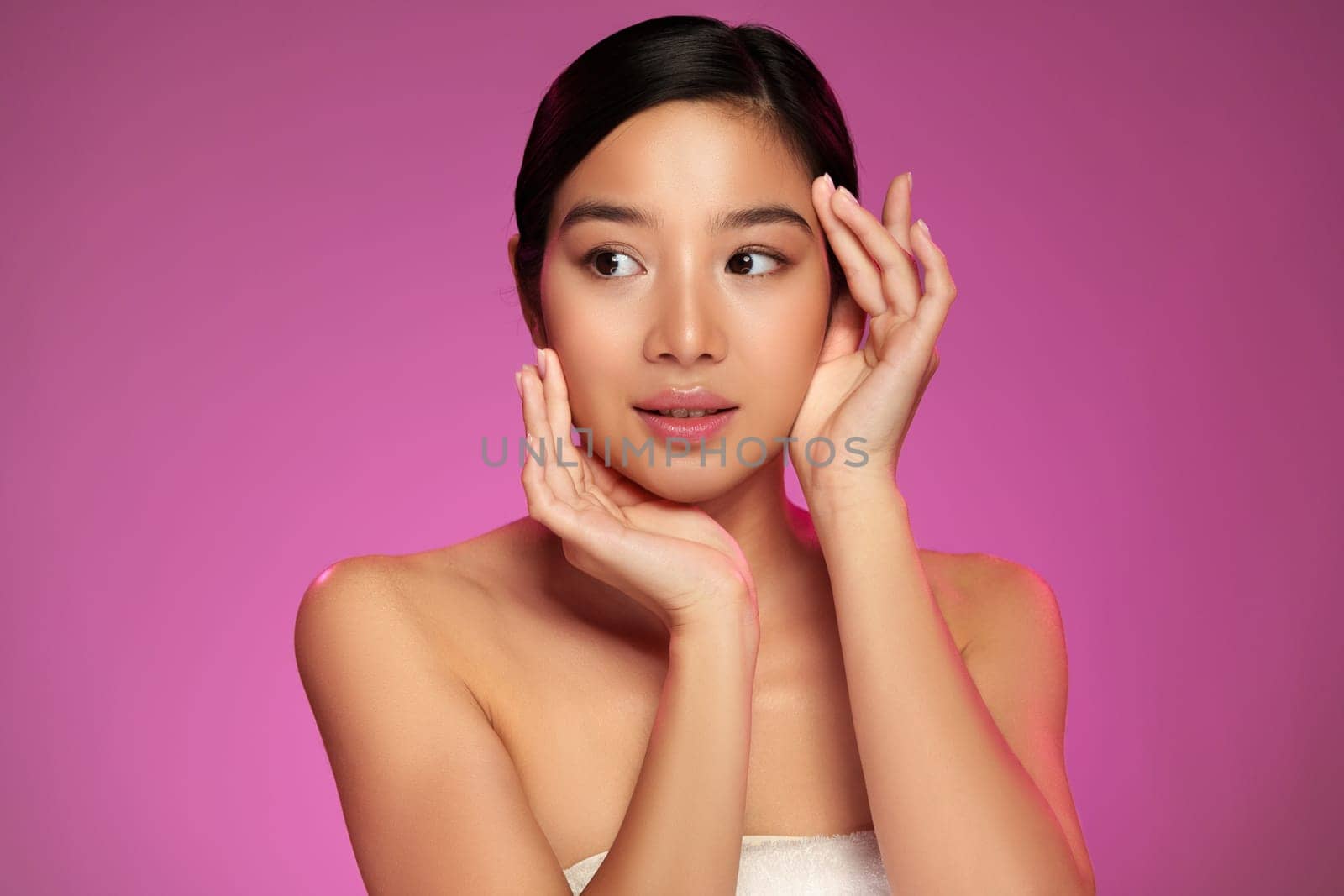 A young woman with clear skin and black hair gently touches her face while showcasing her skincare routine. She poses thoughtfully against a vibrant pink background, radiating confidence and beauty.