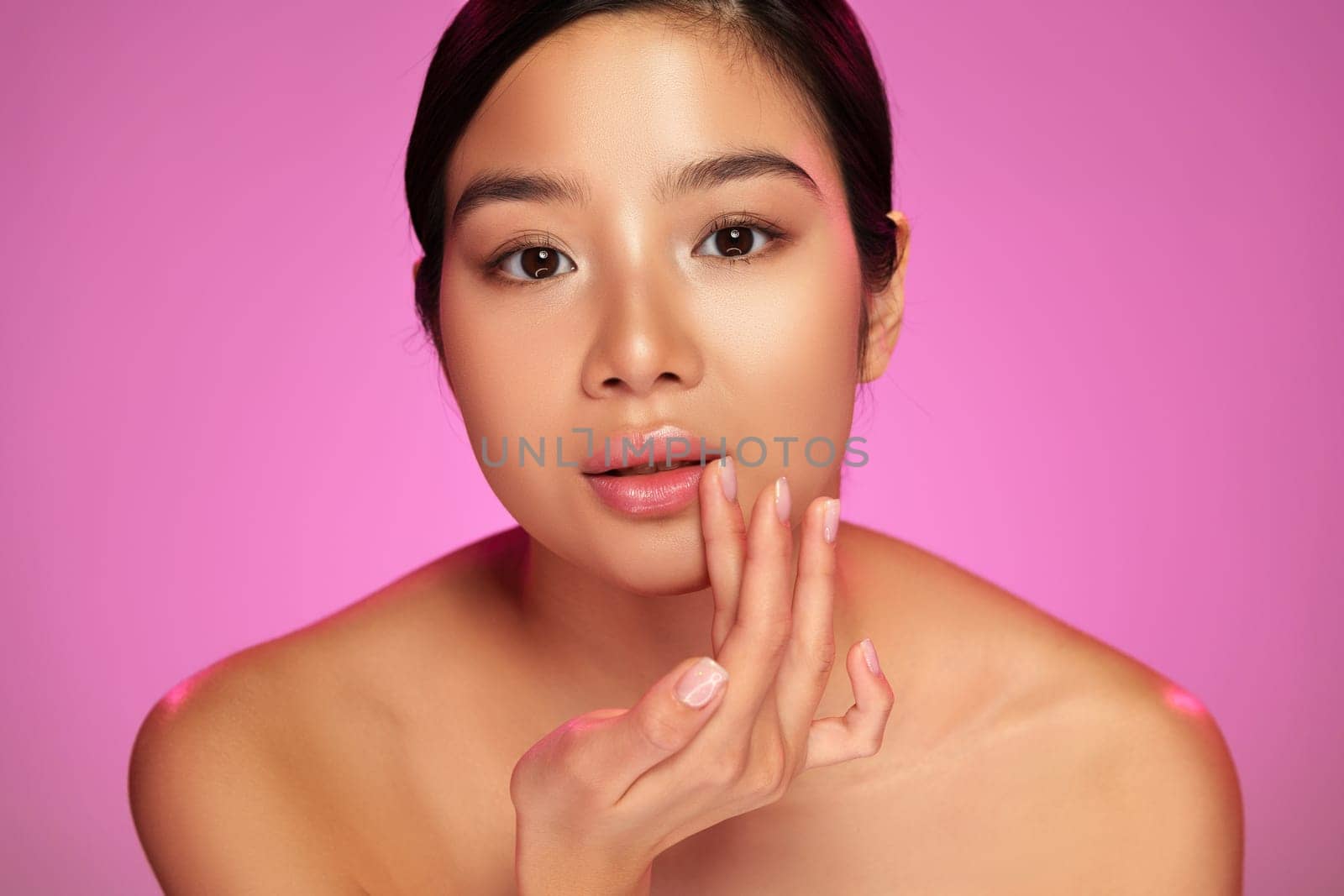 A young model gently touches her face with her hand, showcasing a natural makeup look. The soft pink background enhances her glowing skin and emphasizes beauty and self-care.