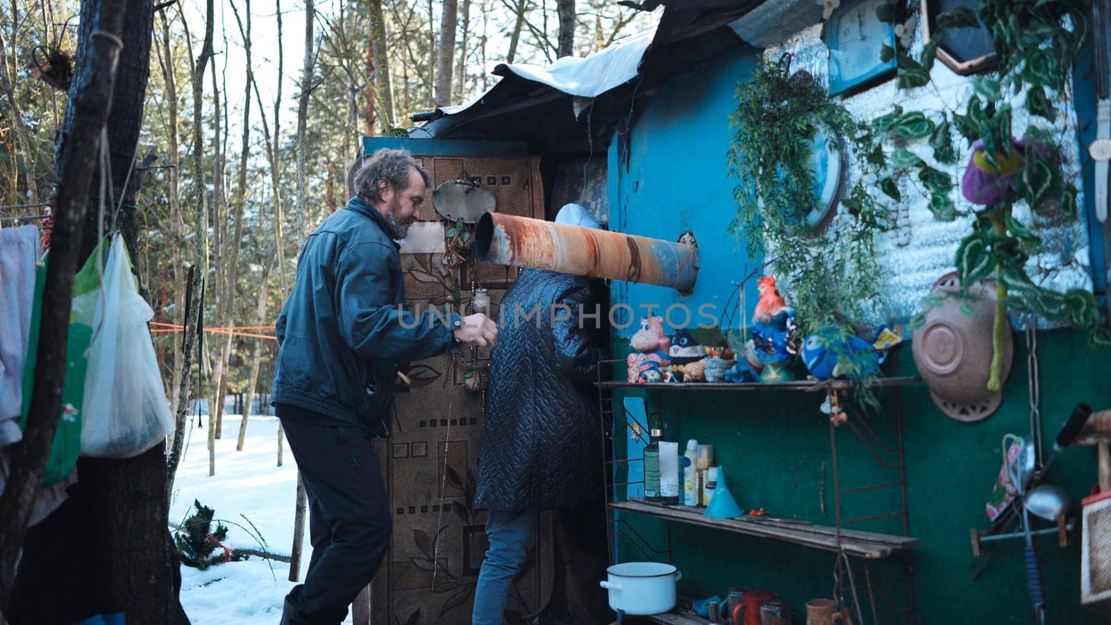 Homeless individuals sheltering in rustic dwelling constructed from salvaged materials, nestled among snow covered winter trees, representing marginalization